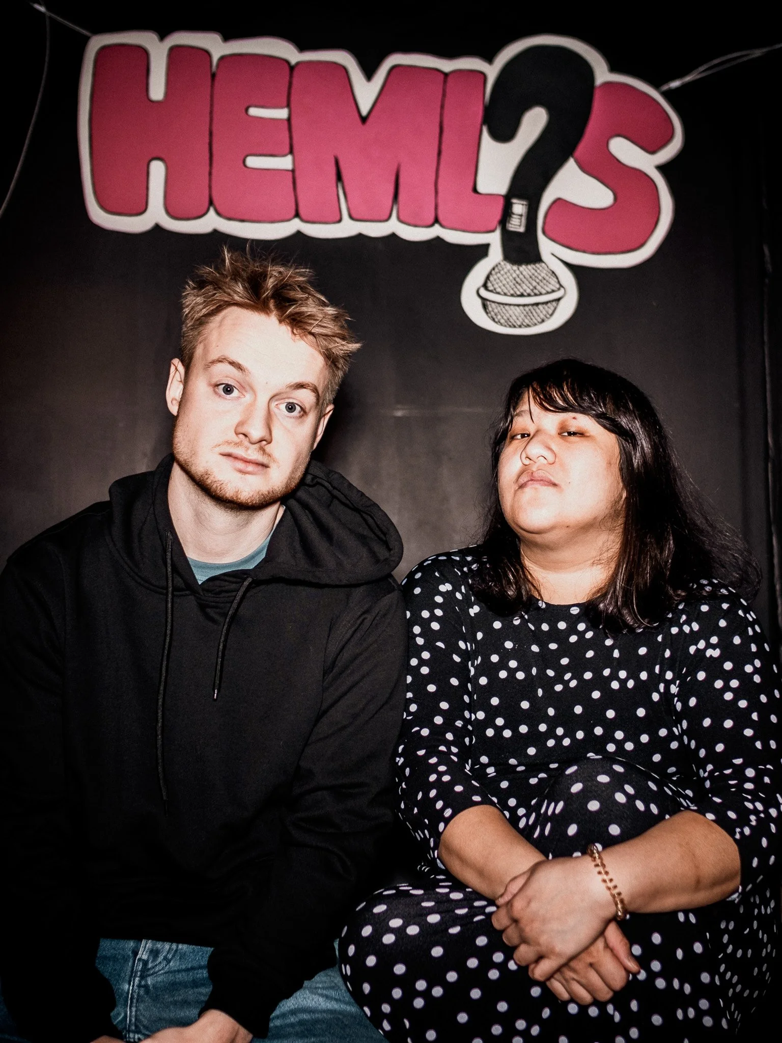 Två personer sitter framför en vägg med texten "HEMLIS" och en mikrofon med en frågetecken ovanför.