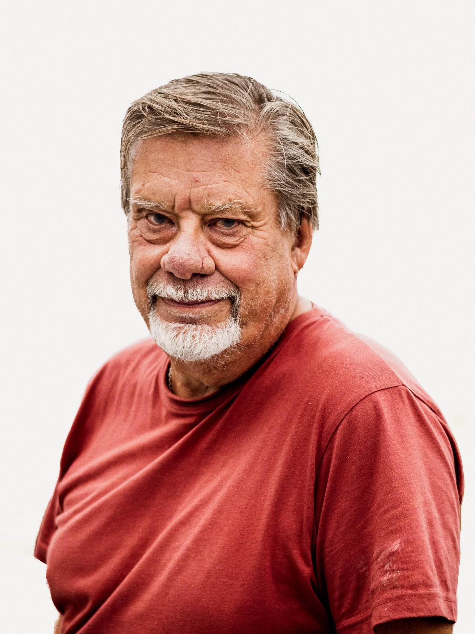 Porträtt av en äldre man med grått hår och skägg, som bär en röd T-shirt, mot en vit bakgrund.
