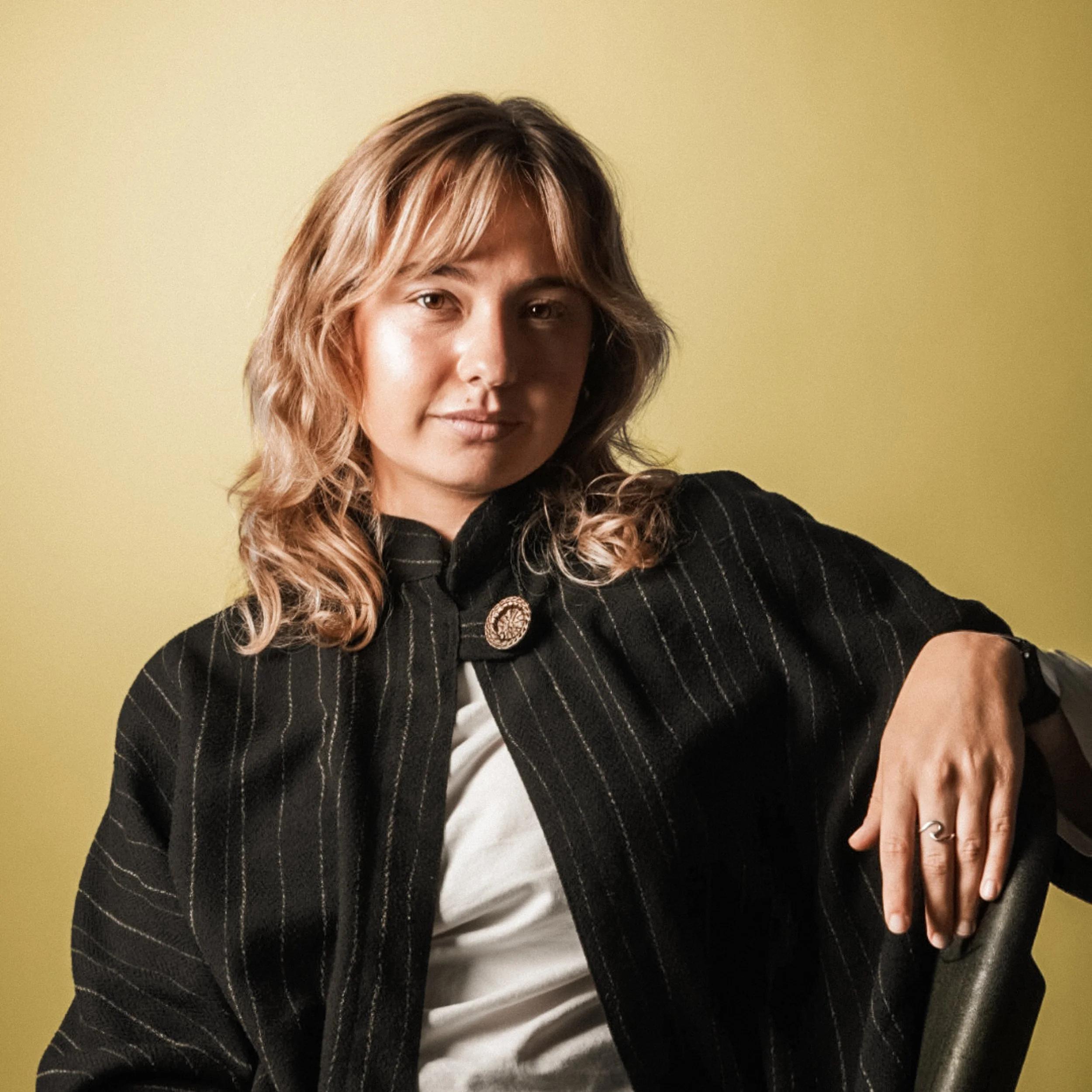 En kvinna med rosa hår som tittar i kameran, sitter i en stol mot en gul bakgrund, bär en svart randig jacka och ett vitt T-shirt.