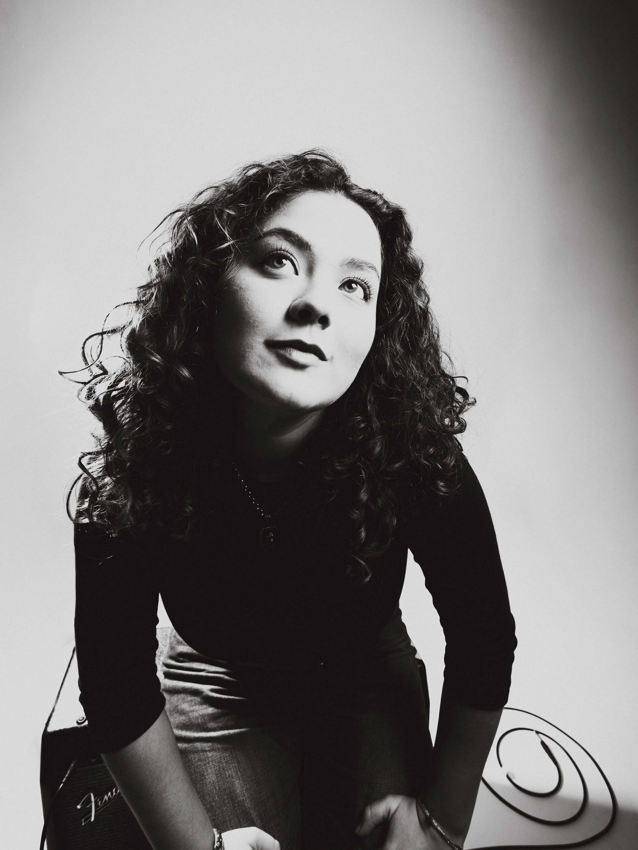 Svartvit bild av en kvinna med lockigt hår, som tittar uppåt, klädd i svart tröja och sittande på en höjd eller stol, med en gitarr i bakgrunden.