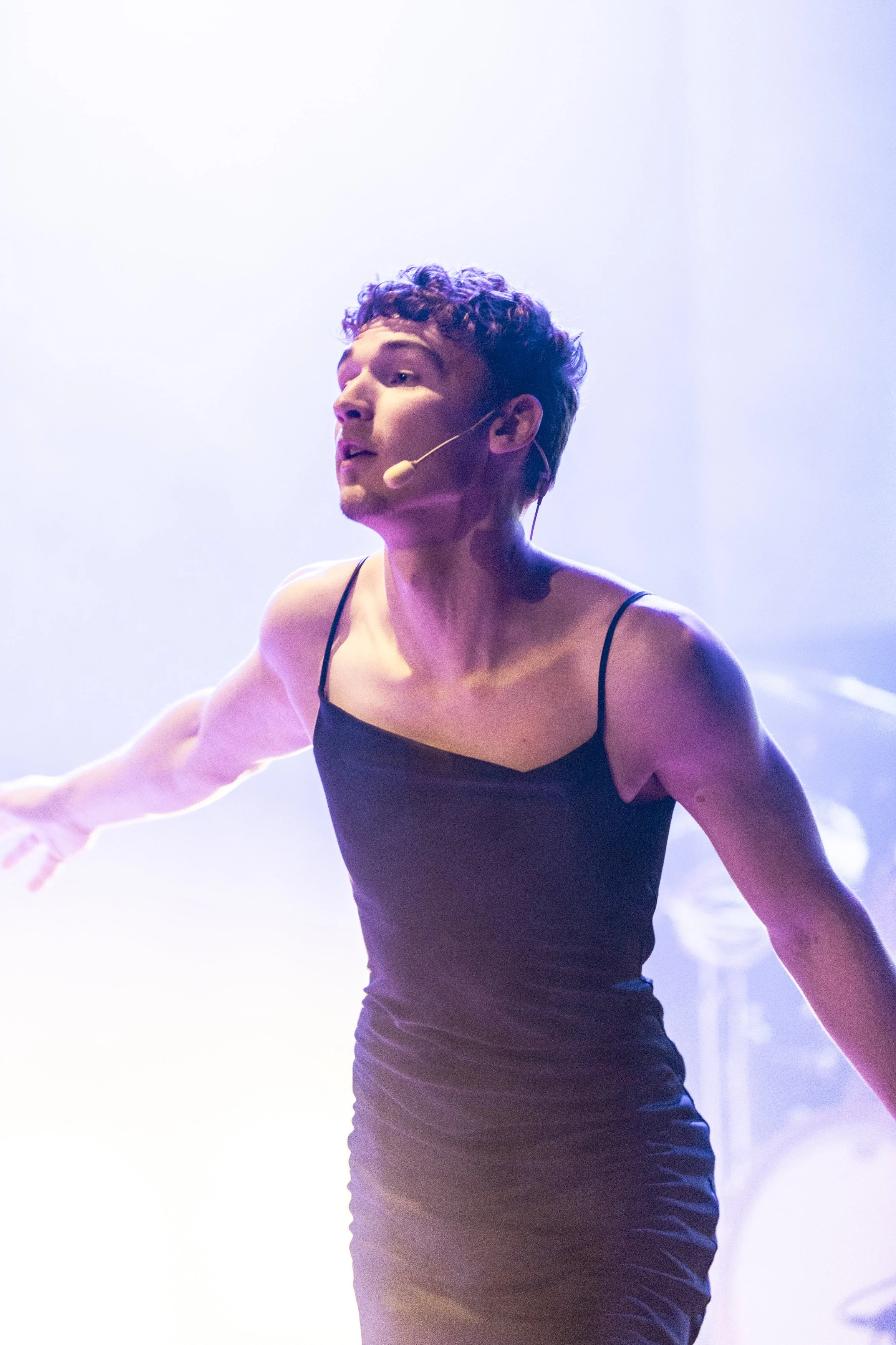 En person som spelar teater på scen, bär en svart klänning med tunna spaghetti-band, med mikrofon på huvudet, under ljust ljus.