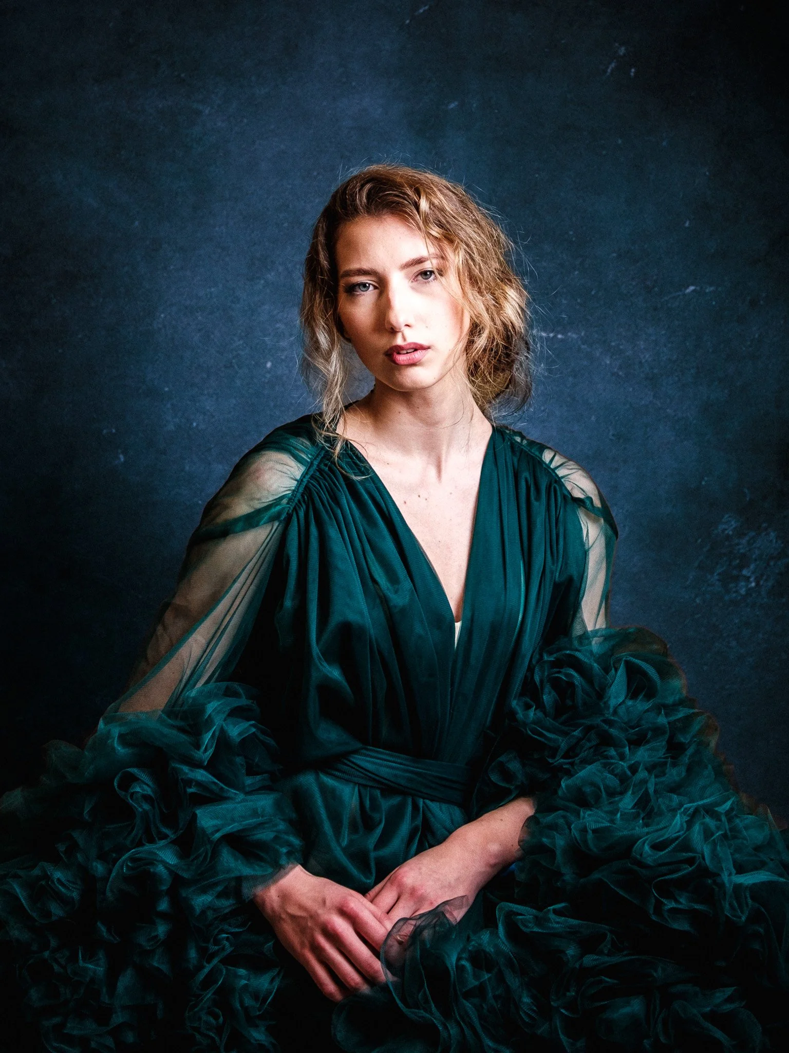 En ung kvinna med lockigt, ljusbrunt hår sitter framför en mörk bakgrund, iklädd en grön, voluminös klänning med rynkig och transparent ärm.