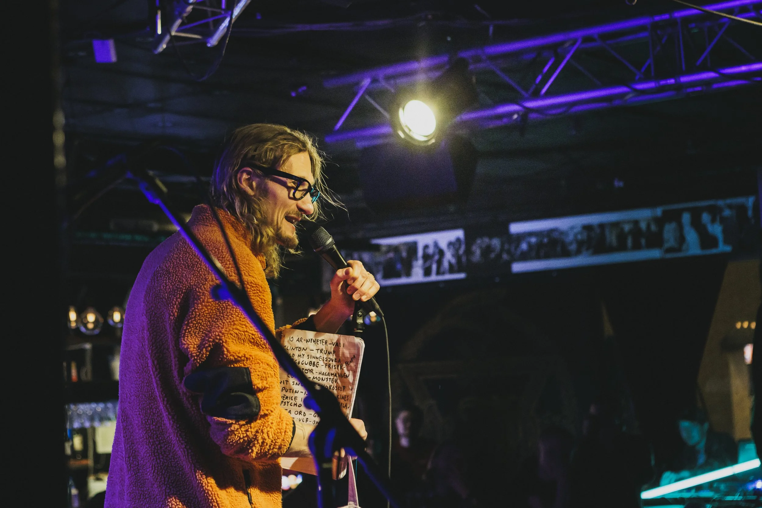 En man med långt hår och glasögon, håller en mikrofon och ett papper, står på scen i en bar eller klubb med mörk bakgrund och blå och lila belysning.