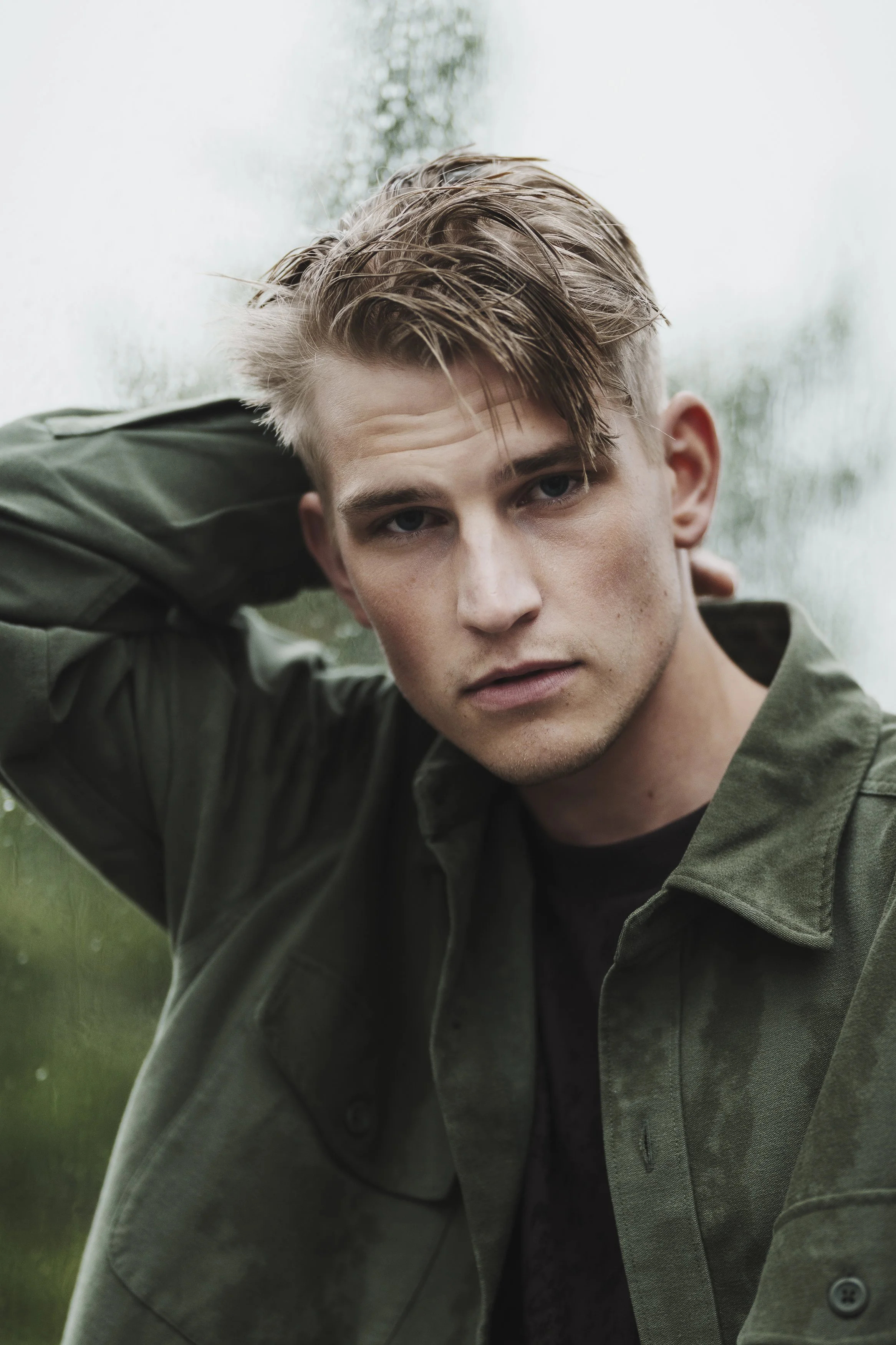 Ung man med blont hår, grönt jacka, står utomhus framför en fönsterruta, tittar in i kameran med en allvarlig min.