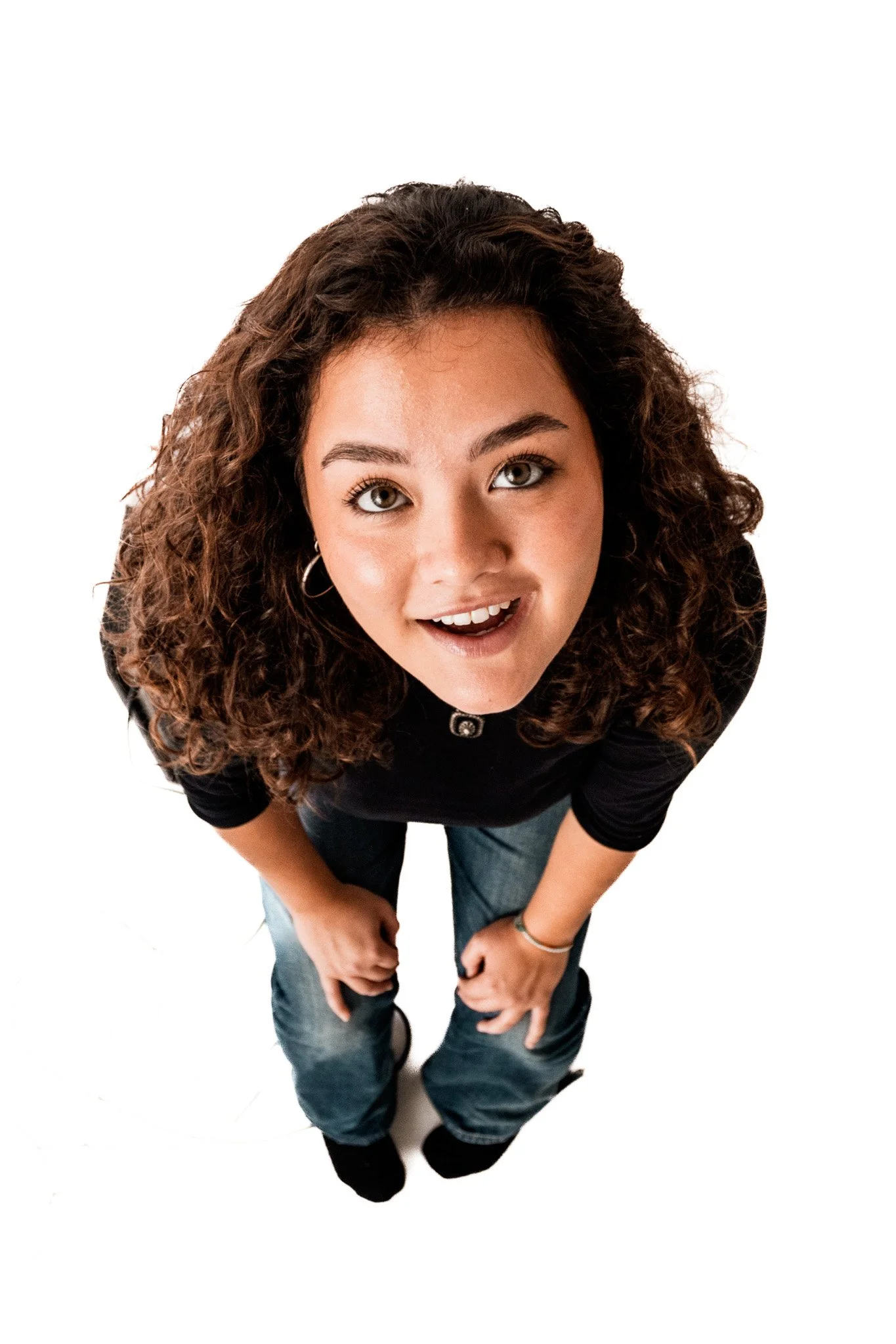 Ung kvinna med lockigt hår, tittar upp mot kameran, ler, iklädd svart T-shirt och jeans, stående mot vit bakgrund.
