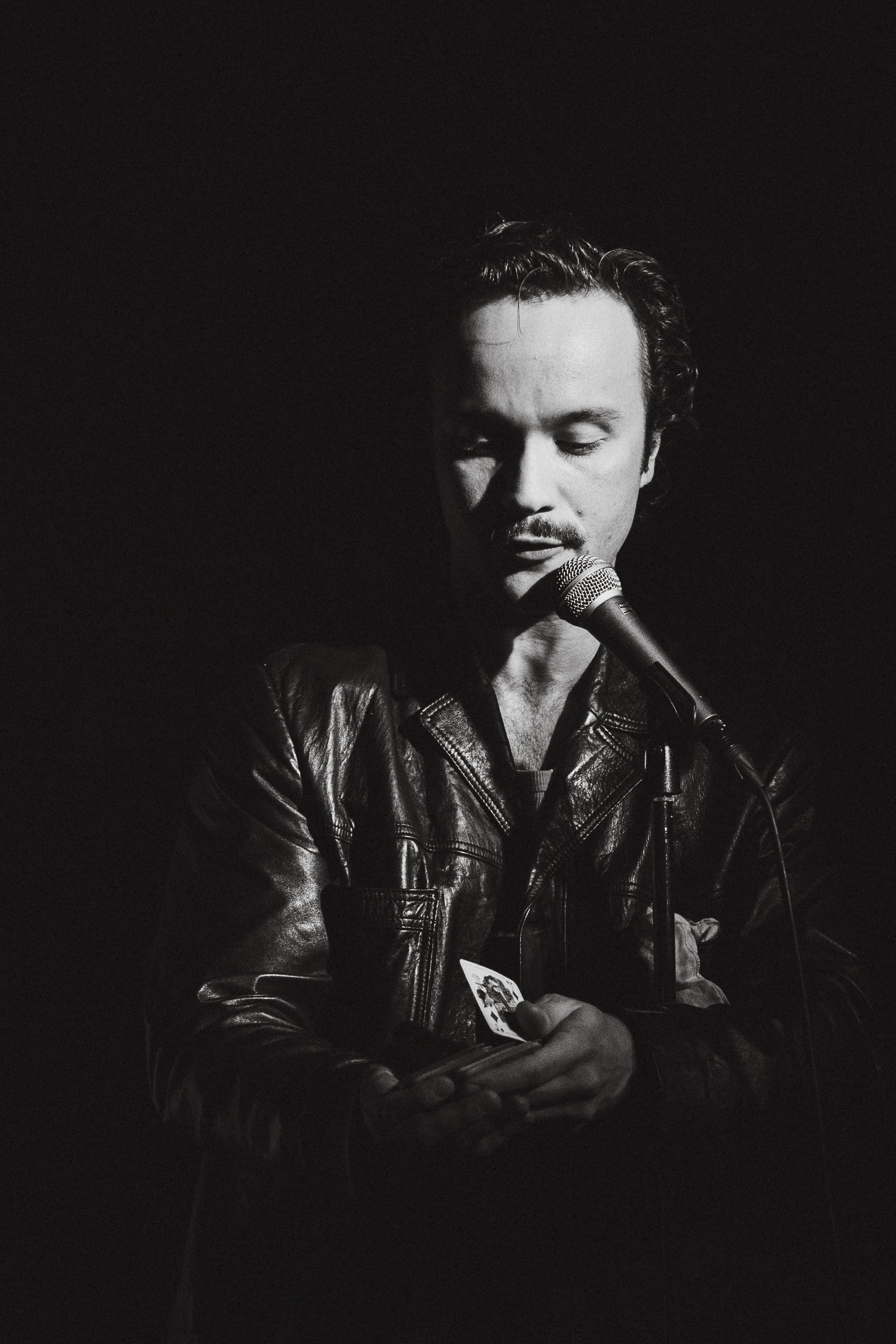 Svartvit bild av en man som står framför en mikrofon, bär läderjacka och håller ett kort i handen. Han har stängt ögon och ett lugnt uttryck.