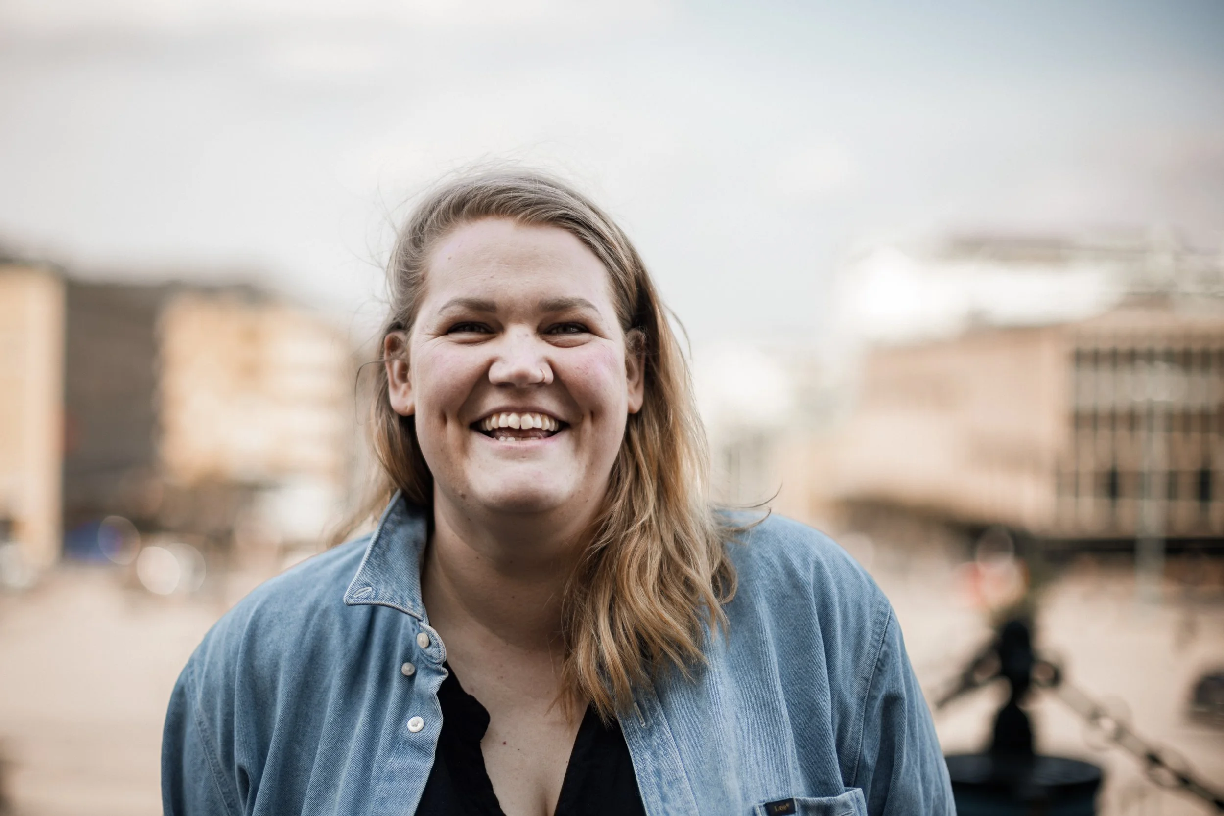 En ung kvinna med långt, lockigt hår, ler och bär en blå jeansjacka, står utomhus i en stadsmiljö under en molnig dag.