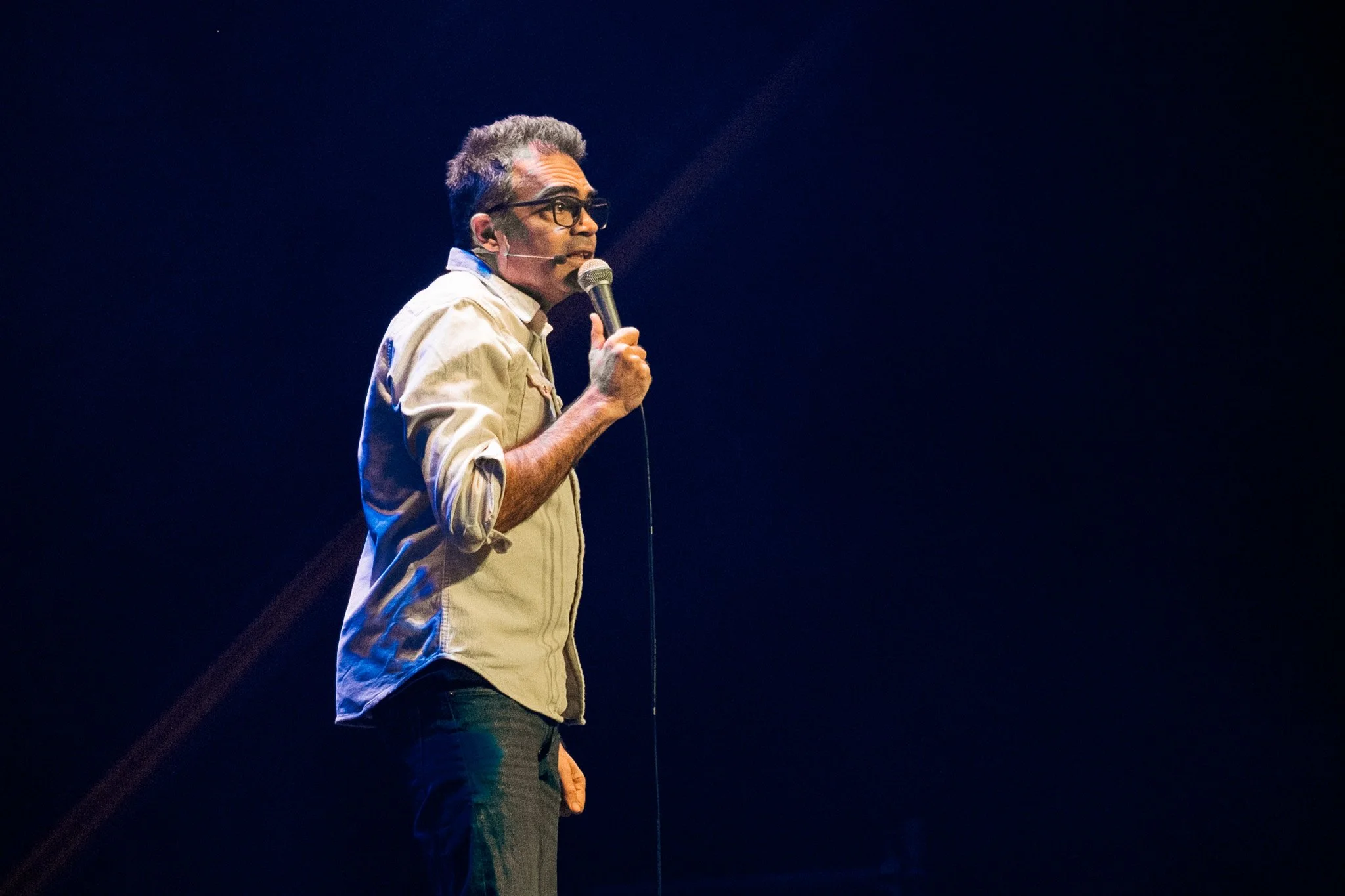 En man med glasögon, mörkt hår och ljus tröja står på scen och talar i mikrofon, med en mörk bakgrund.