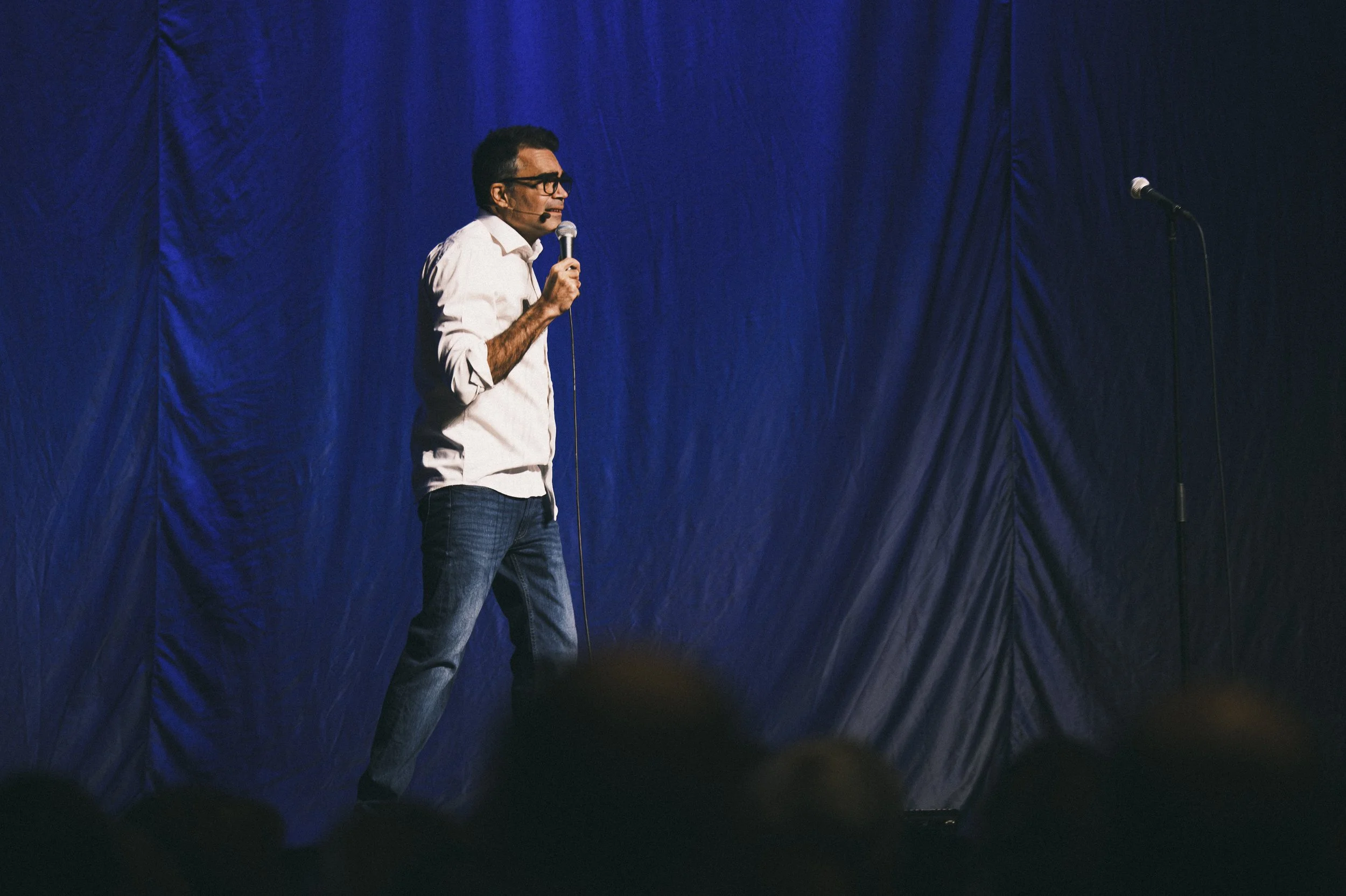 Man står på scen med mikrofon, draperad mörkblå bakgrund, visar stand-up comedy eller föredrag.