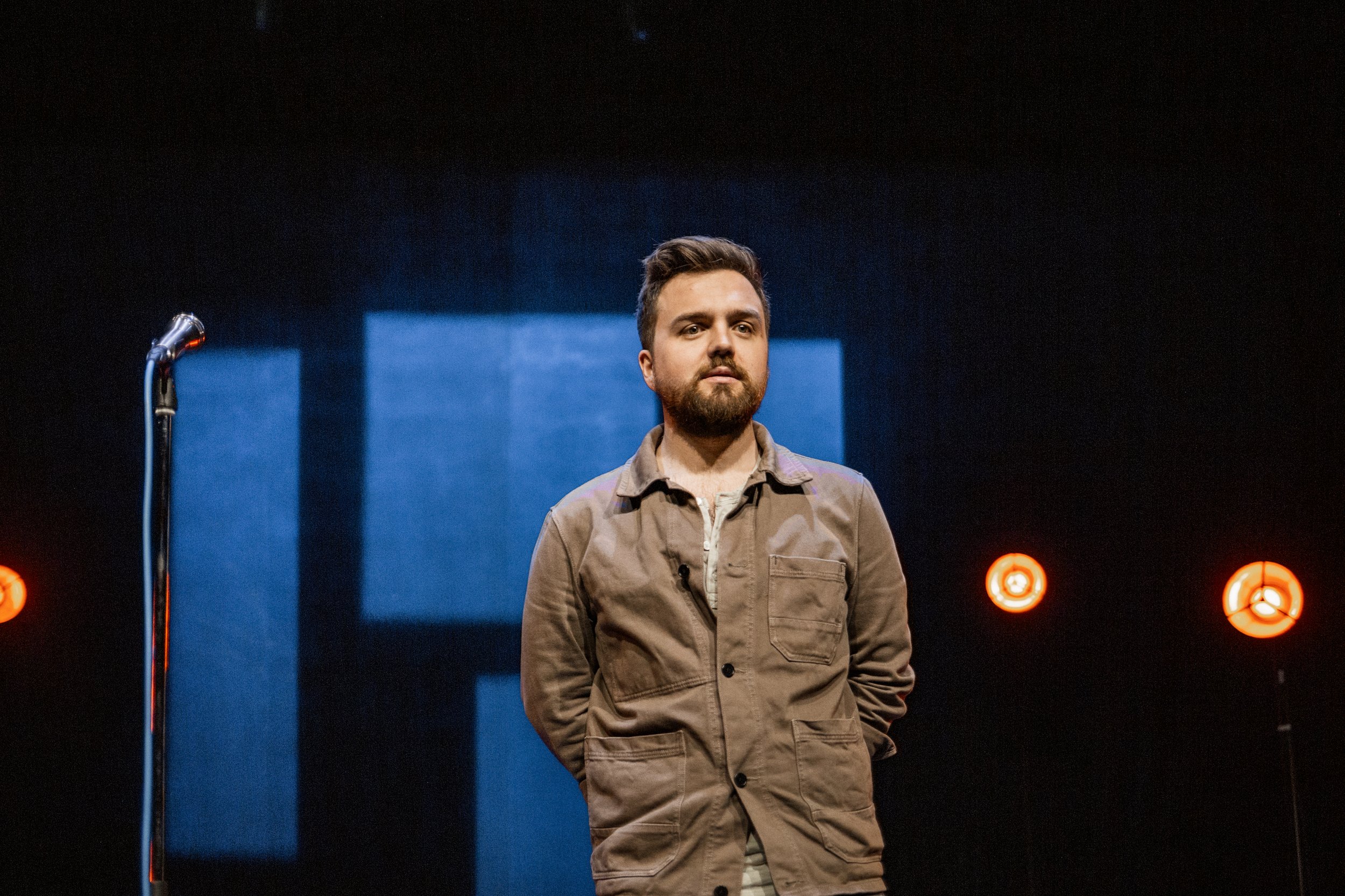 Man står på scen med mikrofon framför sig, bakgrund med blått ljus och tre röda ljus, svart bakgrund.