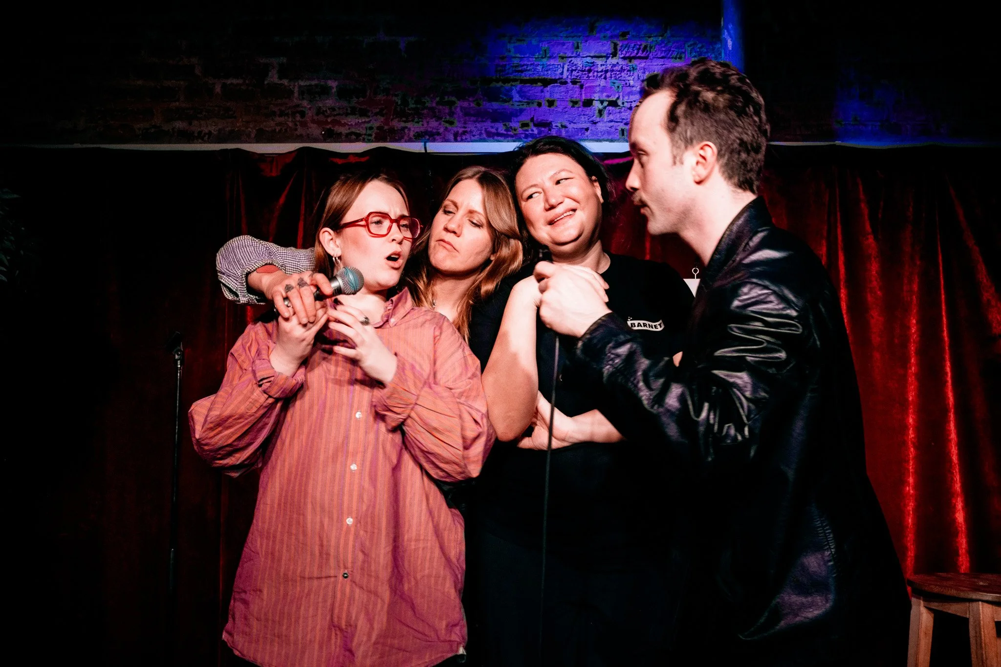 Fyra personer på scen, två kvinnor och en man, står nära varandra och sjunger eller spelar, med ett mörkt röd sammetstyg i bakgrunden.