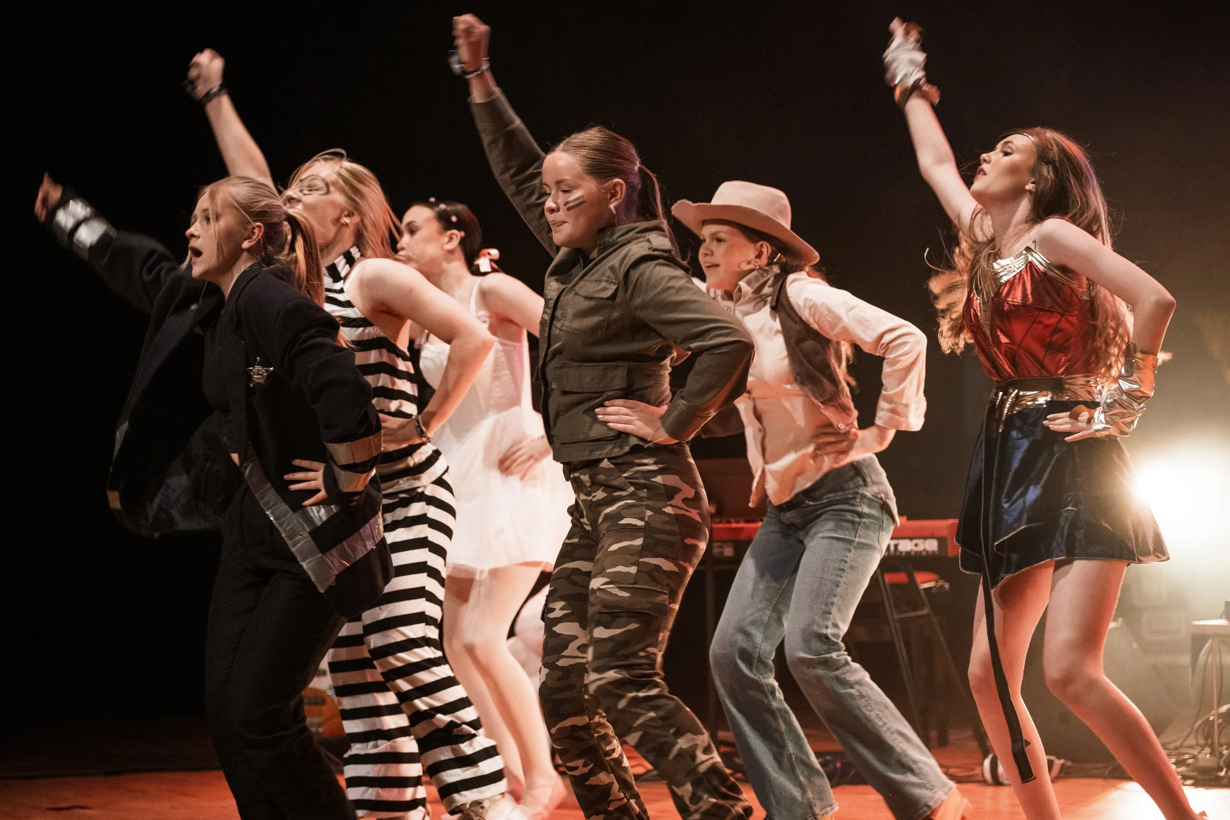 Flera unga kvinnor som dansar på scenen, iklädda olika kläder, i en slumpmässig formation, med ett mörkt bakgrund och spotlight som lyser upp scenen.