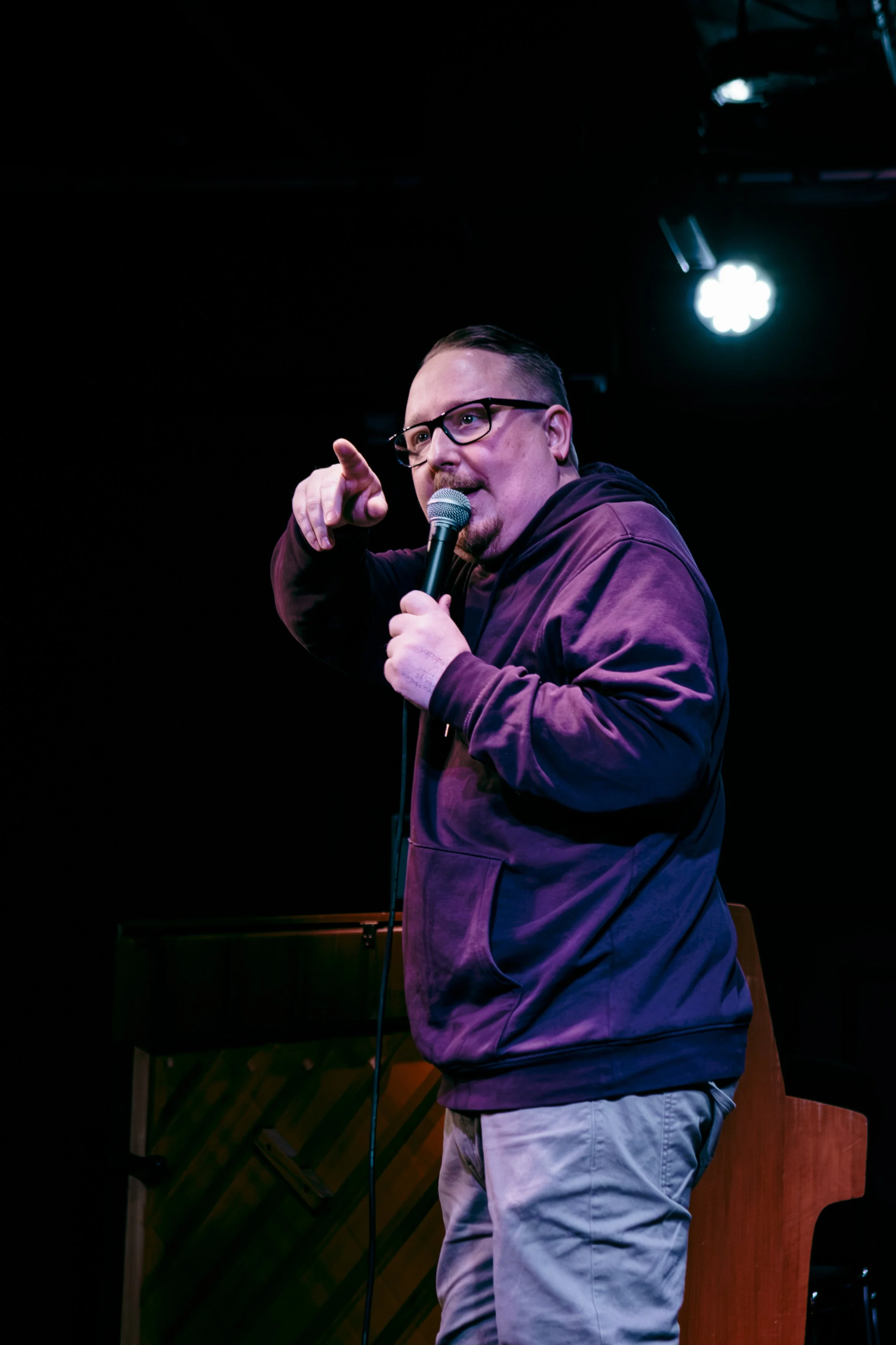 Man med glasögon och skägg, håller en mikrofon och pekar mot kameran, står på en scen med svart bakgrund och ljus.