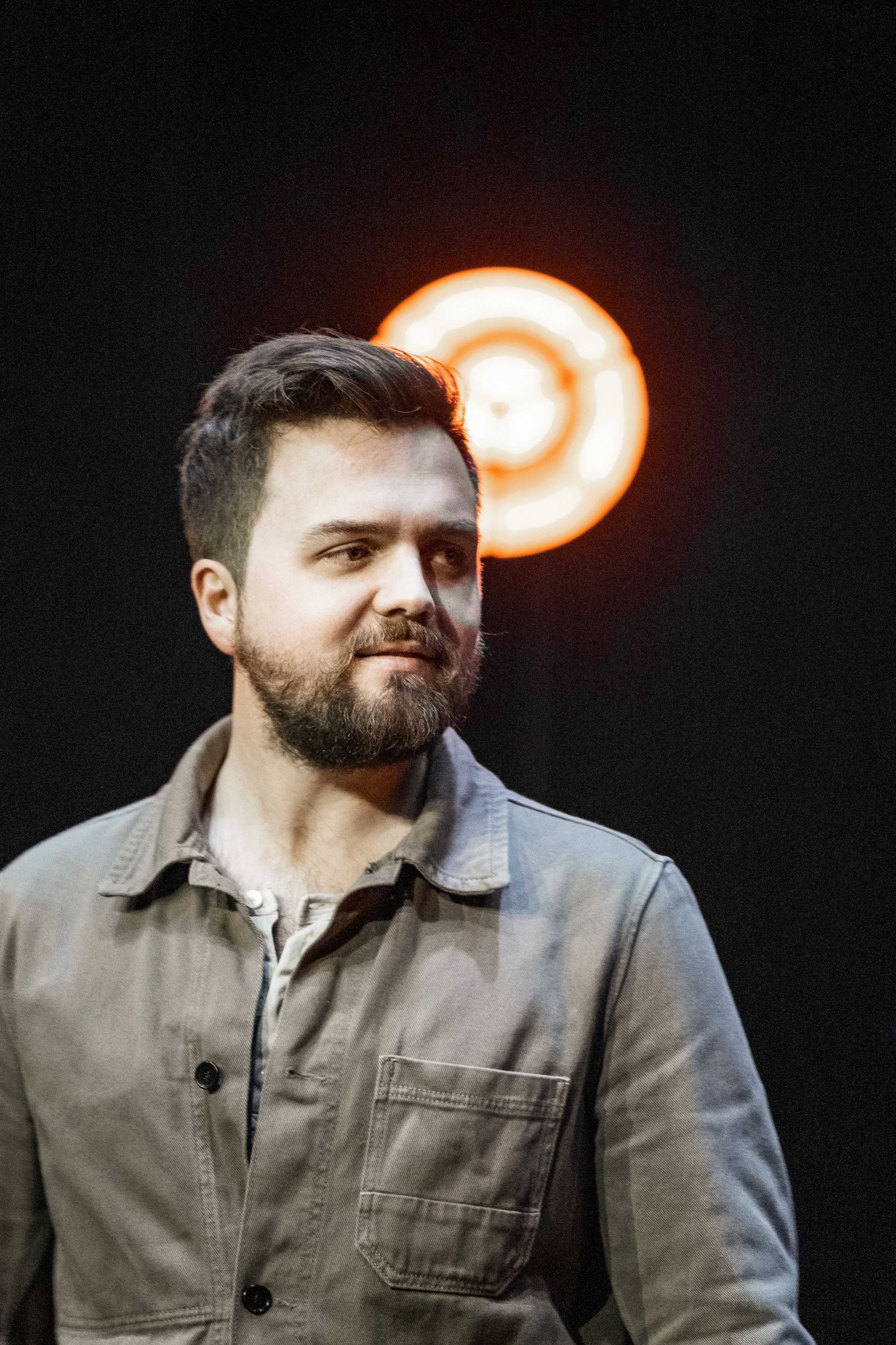 En man med mörkt hår och skägg står framför en svart bakgrund med en röd neonljusskylt bakom sig.