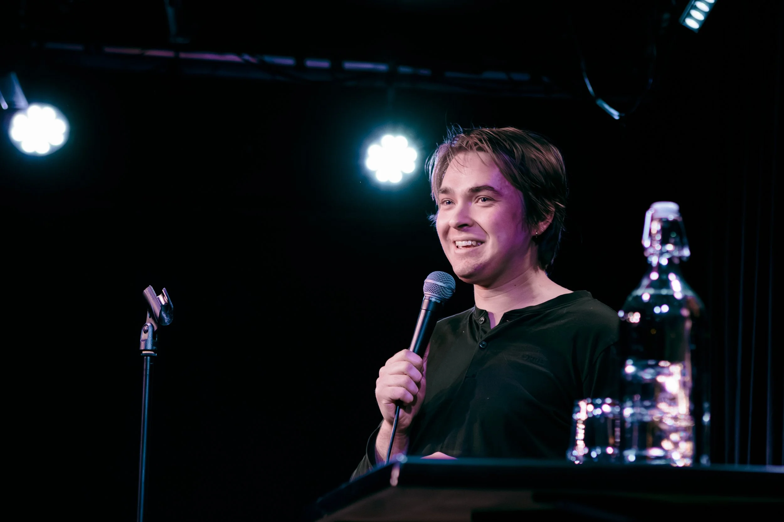 Man står framför en mikrofon på scen, håller den i handen och ler medan han talar. Det finns ett vattenflaska på bordet framför honom och starka stage lights i bakgrunden.