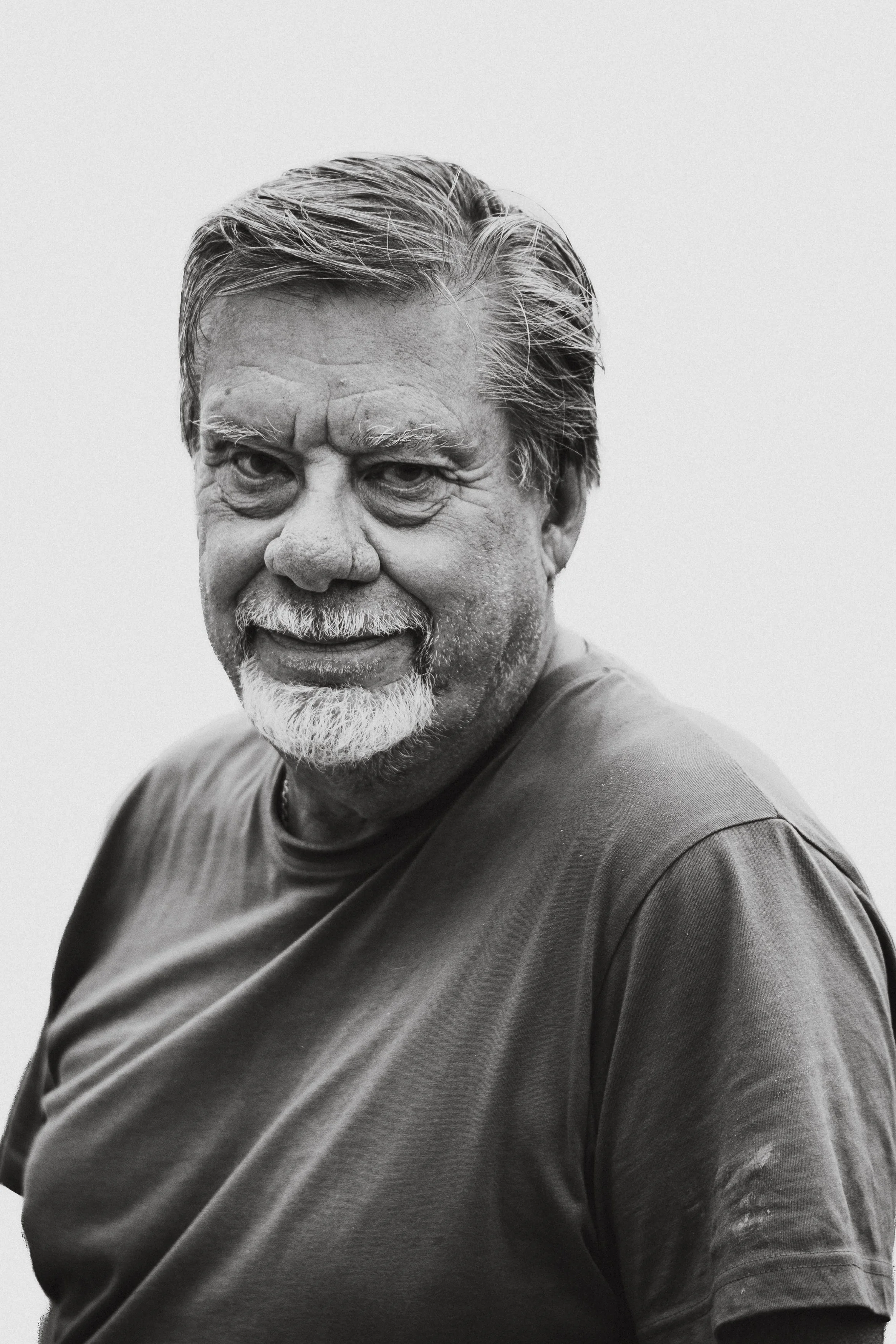 Svartvit bild av en äldre man med grått hår och skägg, klädd i en T-shirt, som ler lite.