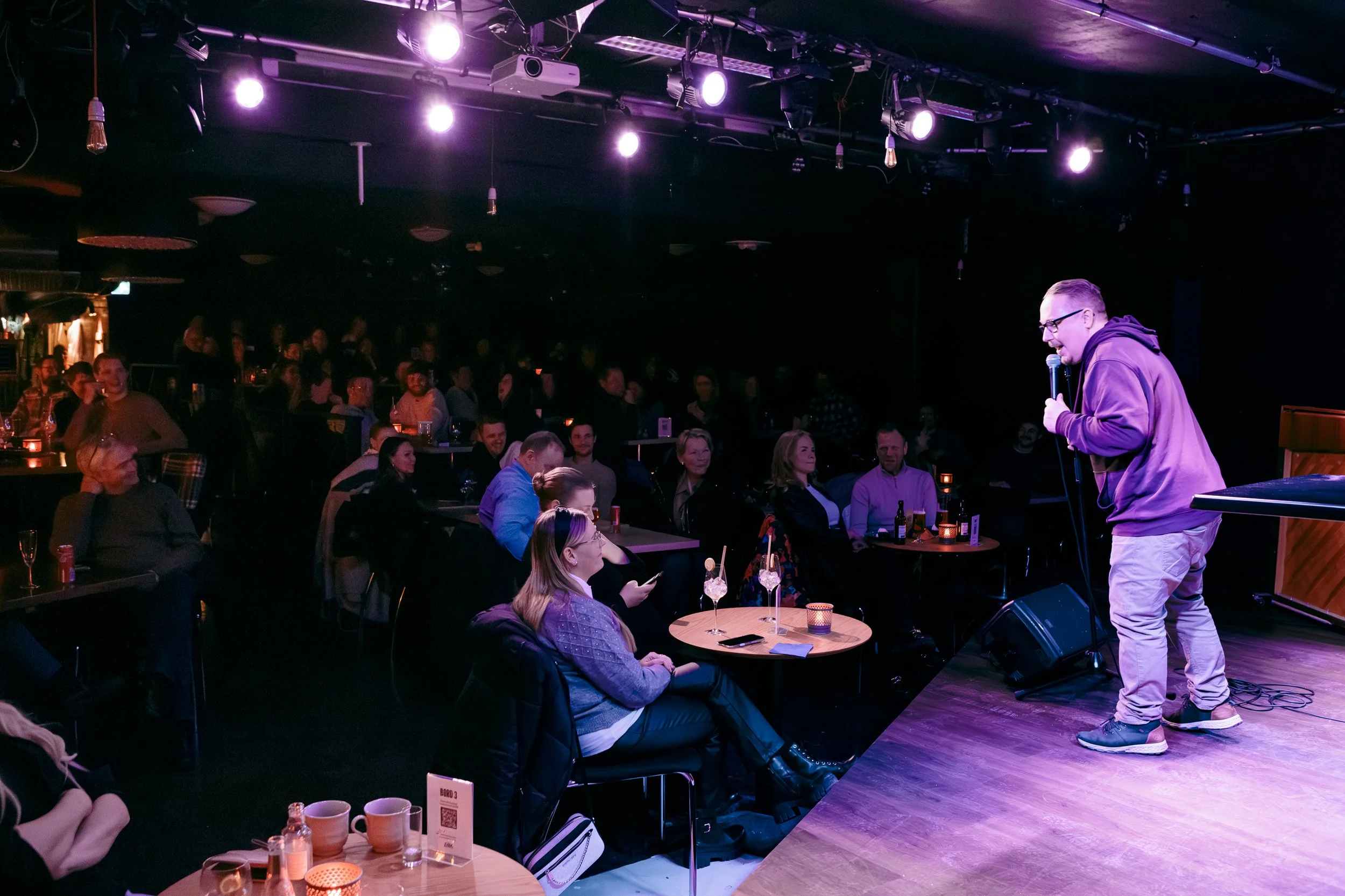 En man står på scenen med mikrofon framför ett publik som sitter vid bord med drycker i en tolkning av en stand-up comedy eller föreställning i en mörk lokal med färgglada ljus.