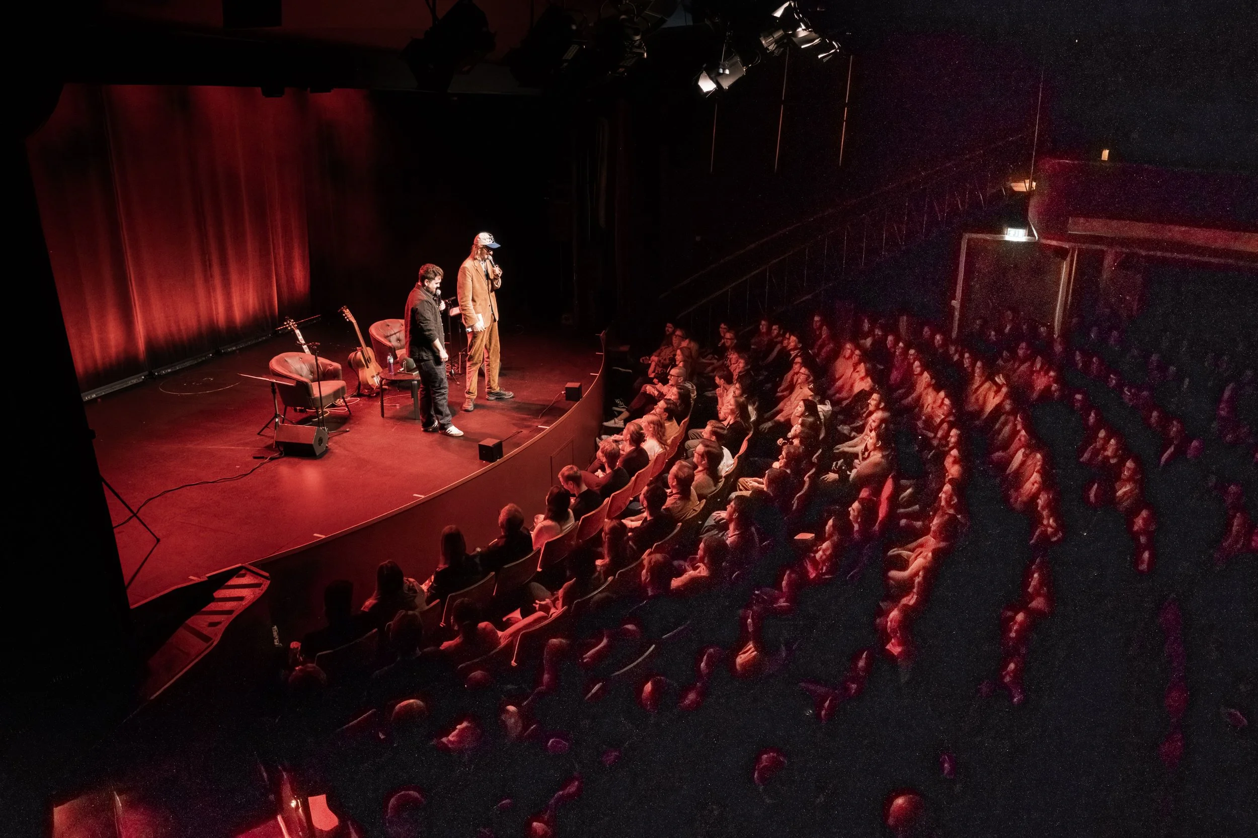 En scen med två personer, en med mikrofon och en med mössa, framför ett mörkt konsertpublikum i en biosal.