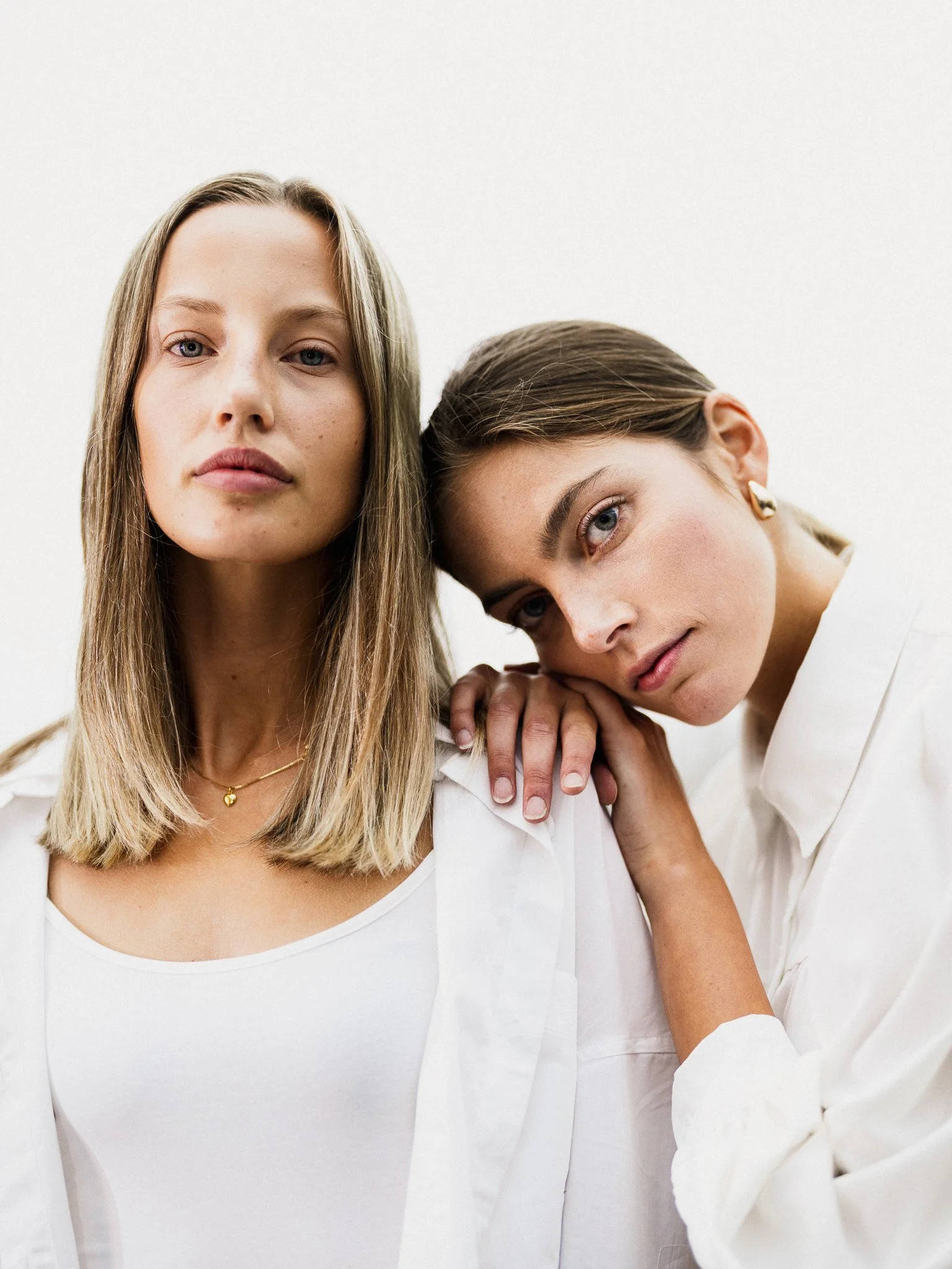 Två unga kvinnor i vita kläder, den ena står blickstilla och den andra lutar huvudet mot hennes axel, båda ser in i kameran.