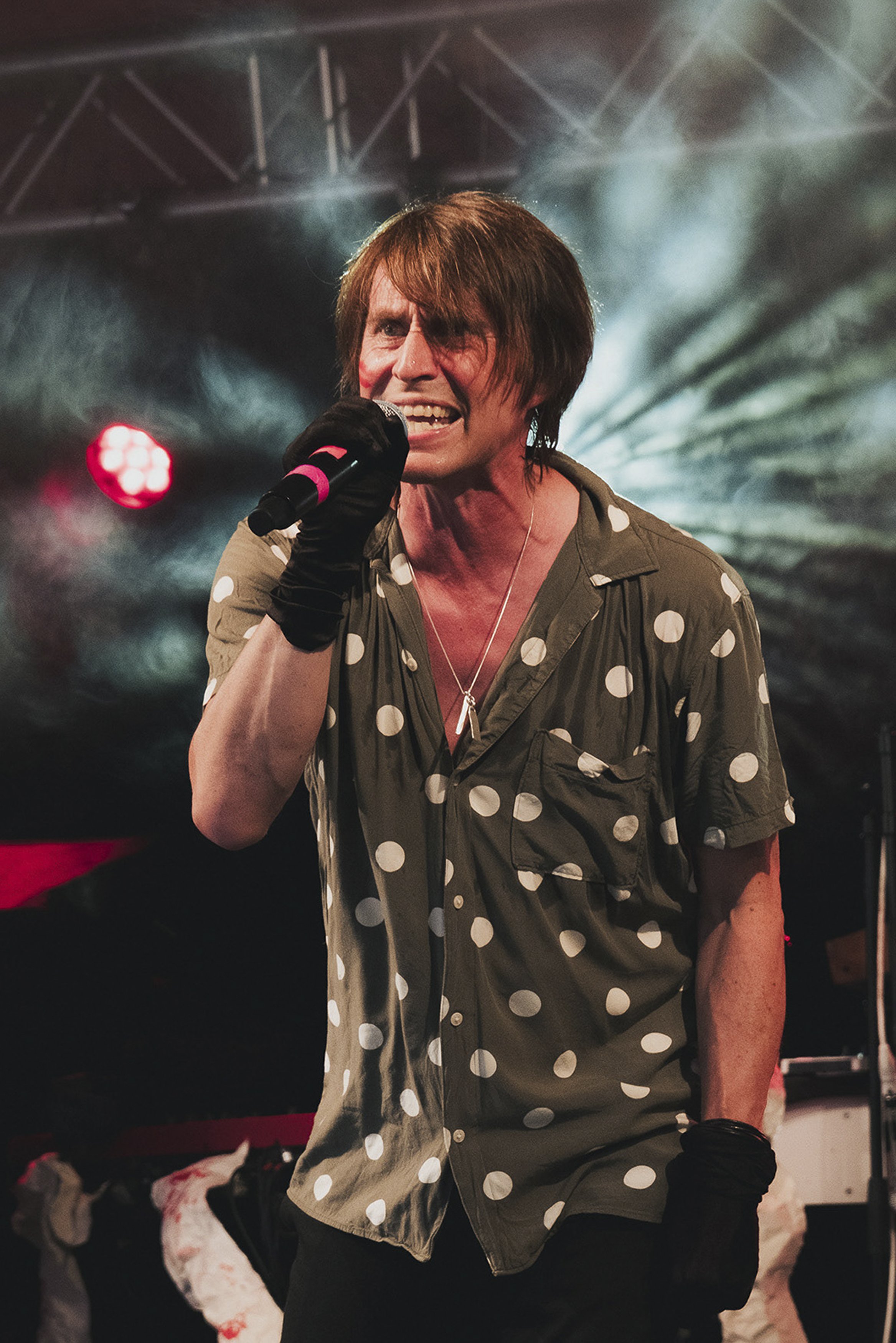 En man med brunt hår som sjunger på scen, bär en grön skjorta med vita prickar och svarta handskar, med scenljus i bakgrunden.