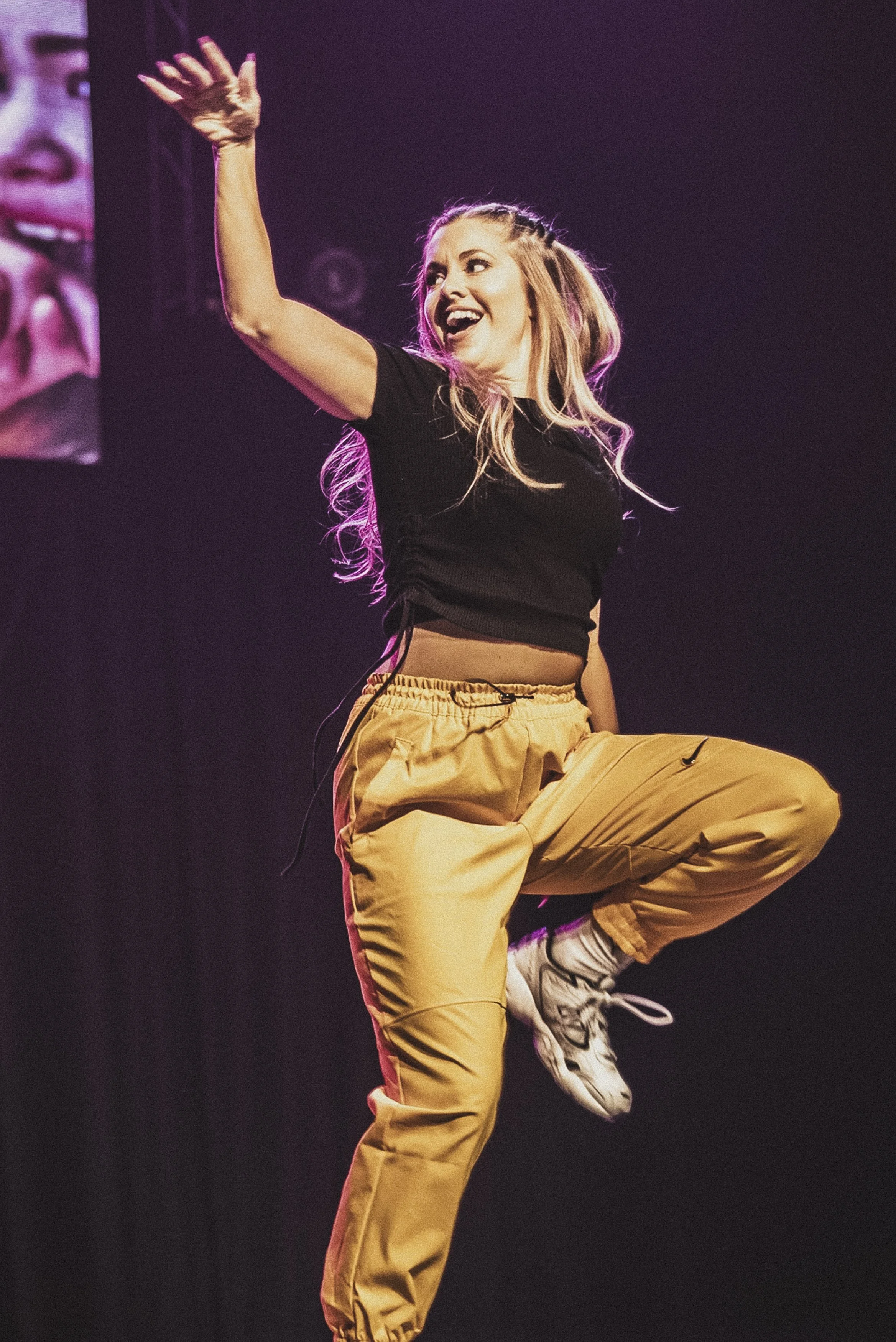 En kvinna dansar på scenen, klädd i svart t-shirt och gulaktiga byxor, med ett brett leende och en arm i luften.