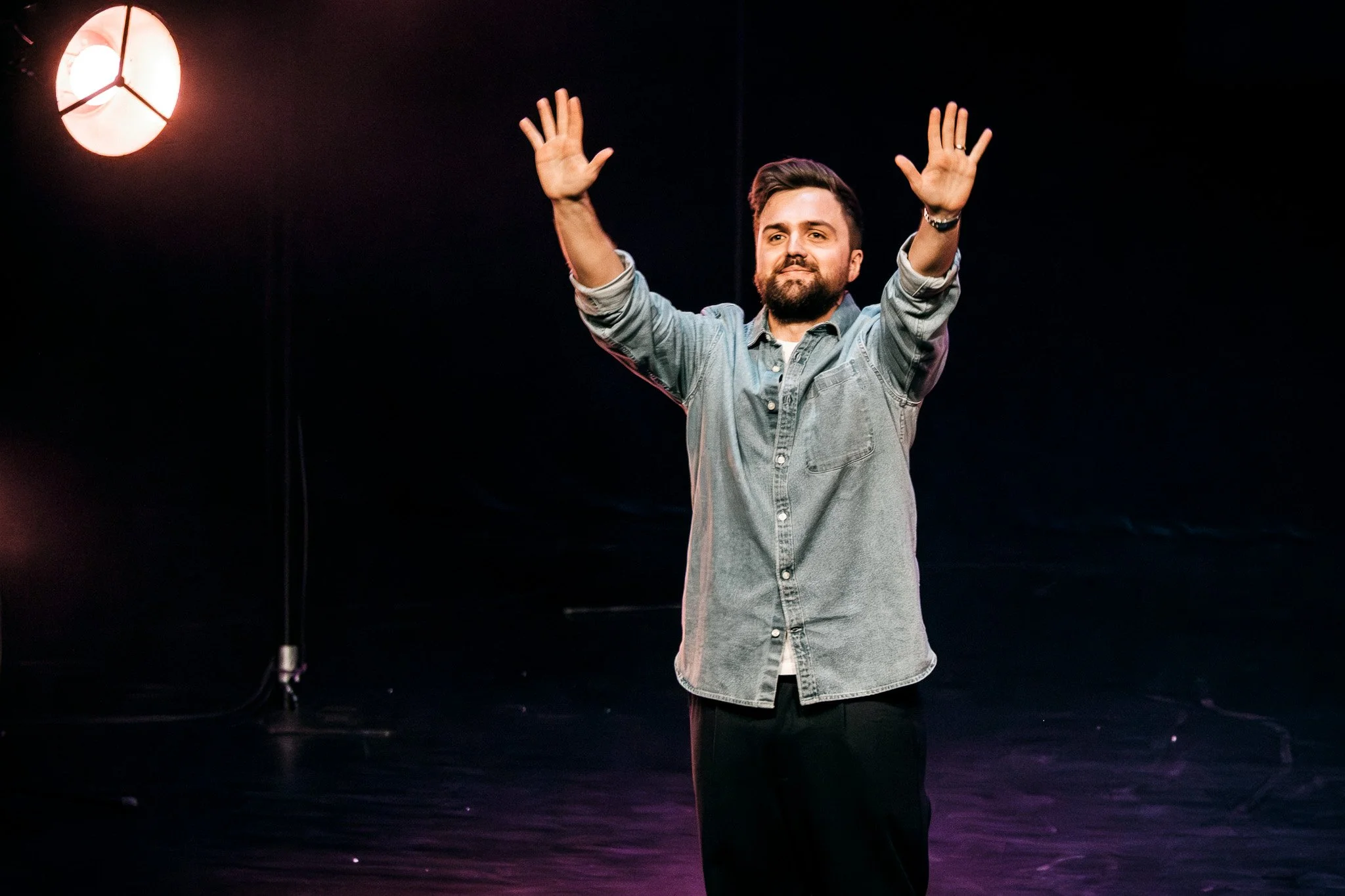 Man står på scen med händerna i luften, mörk bakgrund, scenbelysning