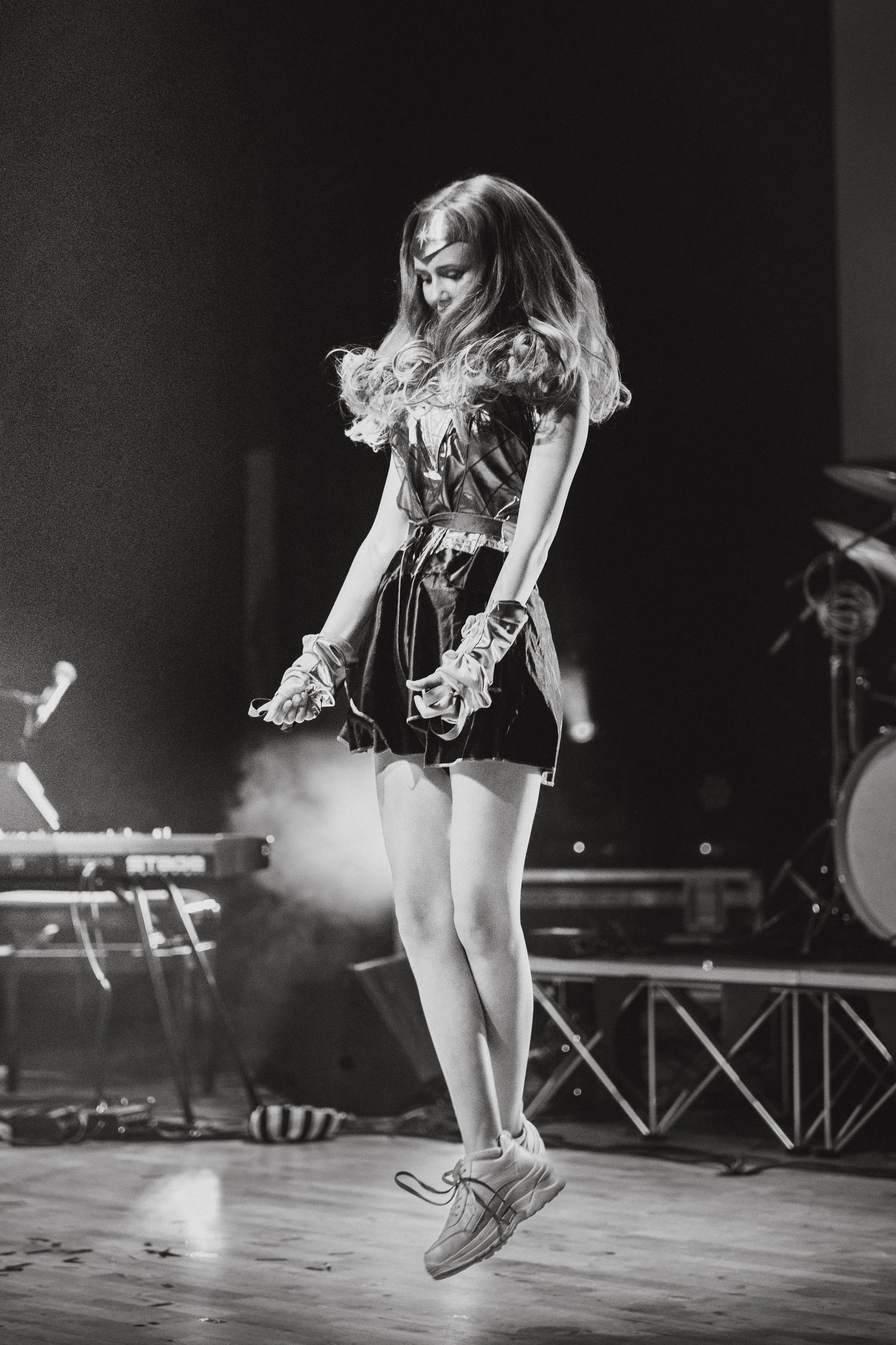 En svartvit bild av en ung kvinna iklädd en svart klänning och sneakers som hoppar i luften på en scen med musikutrustning runt omkring.