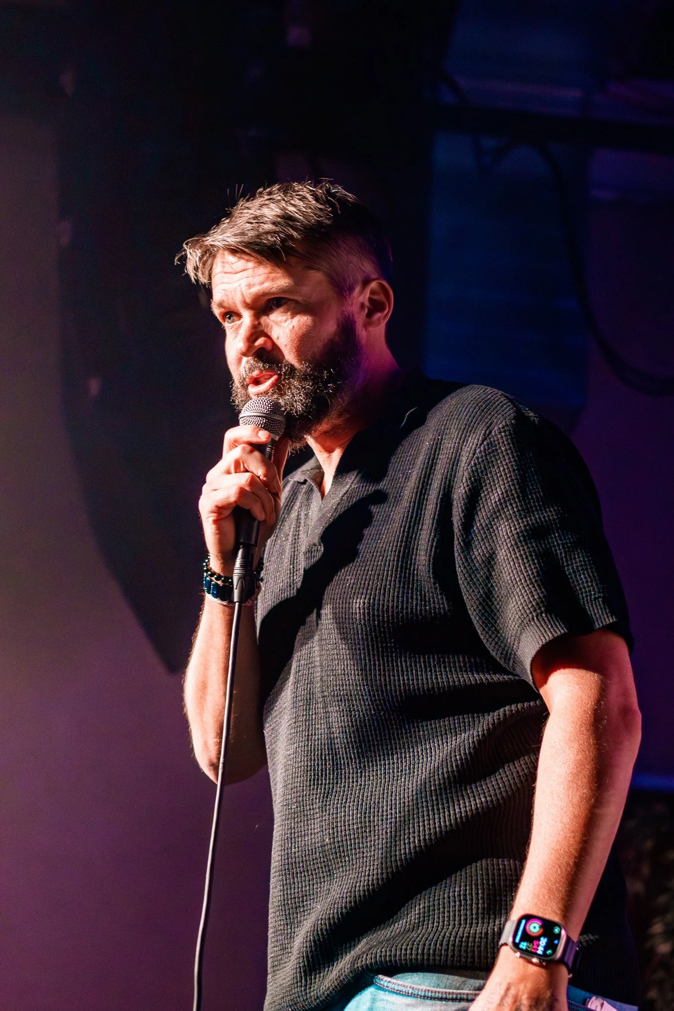Man med skägg och kort hår håller mikrofon på scen, belysning i bakgrunden.