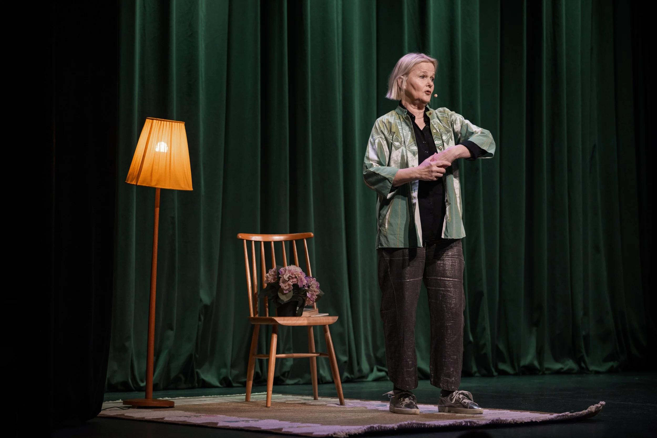 En äldre kvinna står på en scen med gröna gardiner i bakgrunden. Hon är klädd i en luftig jacka och mörka byxor, och hon talar eller gestikulerar. Det finns en golvlampa med orange lammskärm, en trästol med en blomkruka med rosa blommor, och ett lite