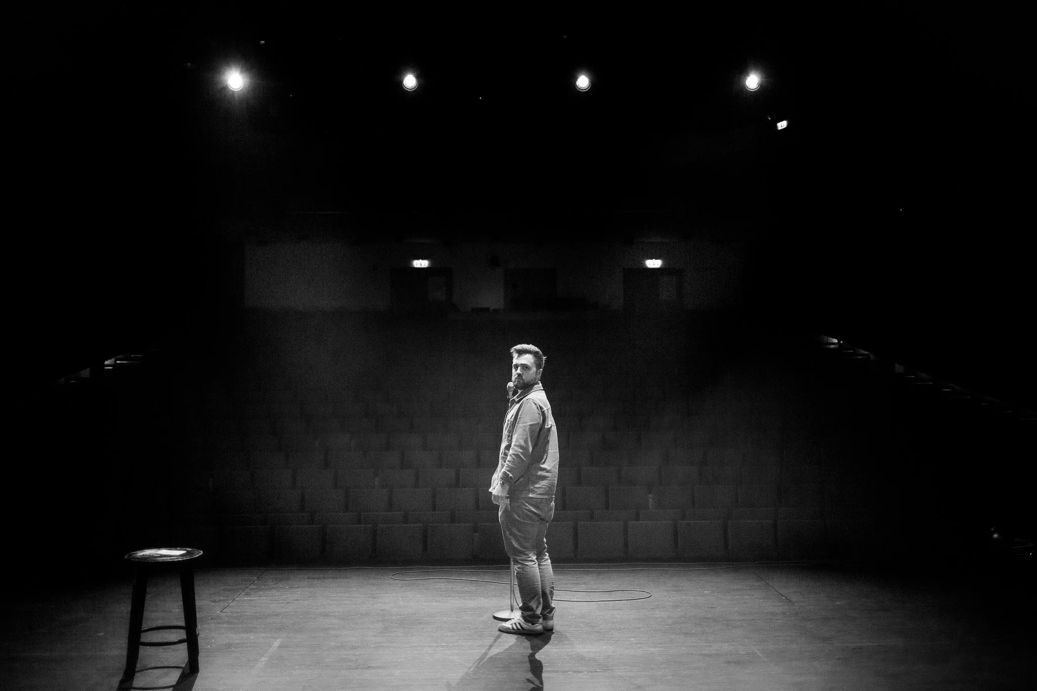 En man i jeans och hoodie står ensam på en tom scen med en stol till vänster och ett mikrofonstativ framför honom, i ett mörkt teatersal.