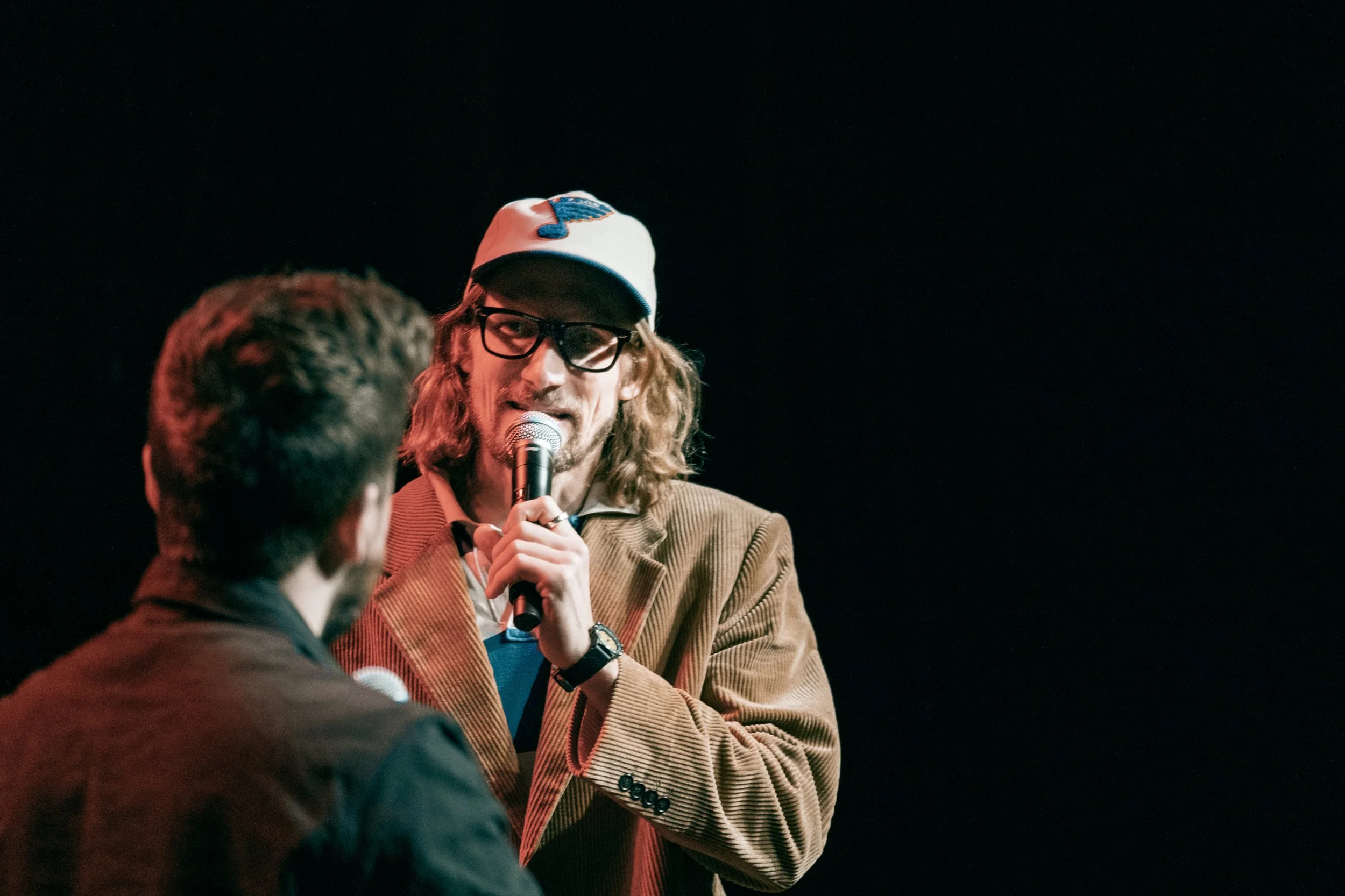 En man med långt lockigt hår, glasögon och hatt, talar i mikrofon under en scen, tittar på en annan person.