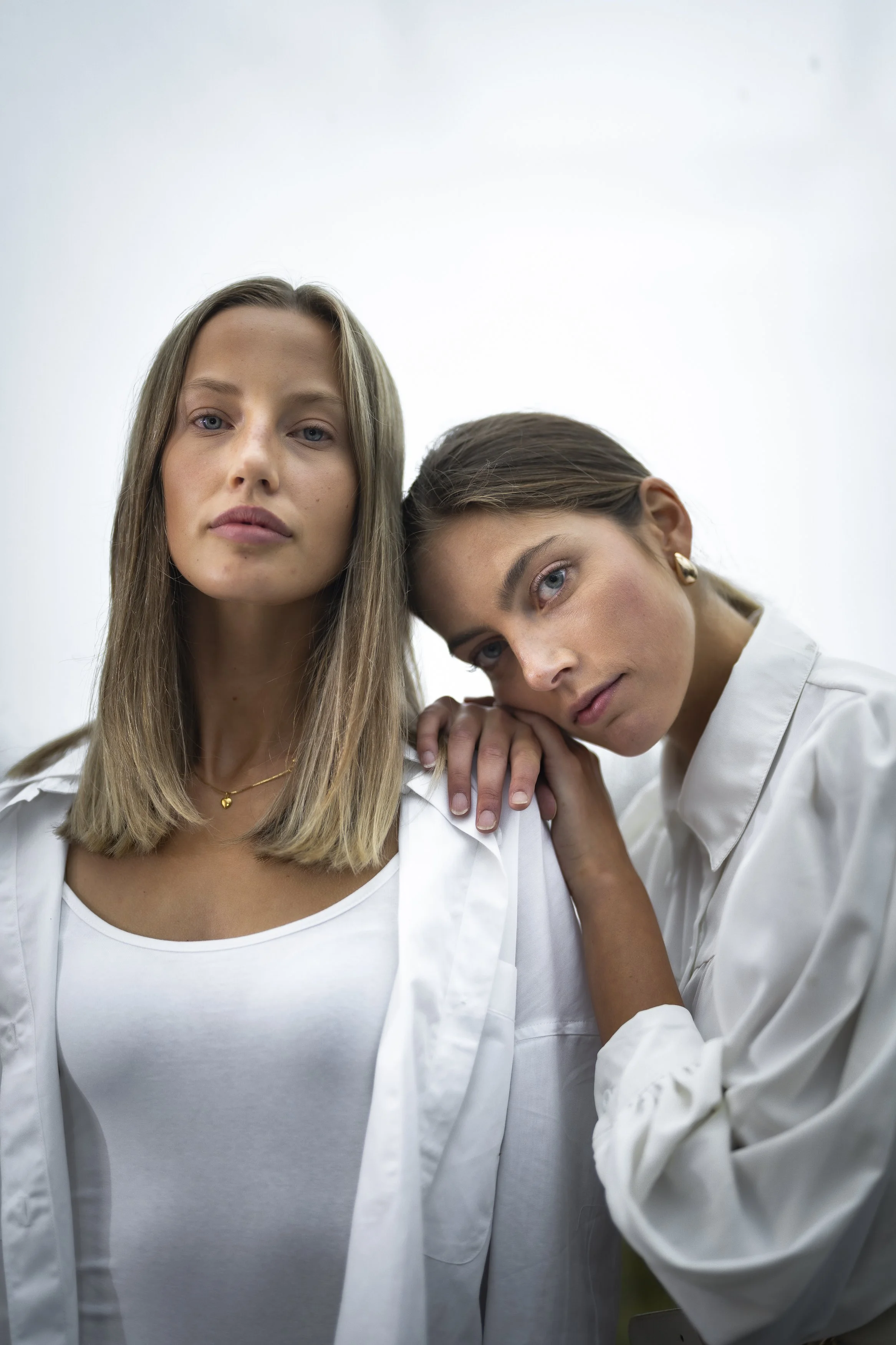 Två unga kvinnor i vita kläder, den ena vilar sitt huvud på den andra's axel, mot en neutral bakgrund.
