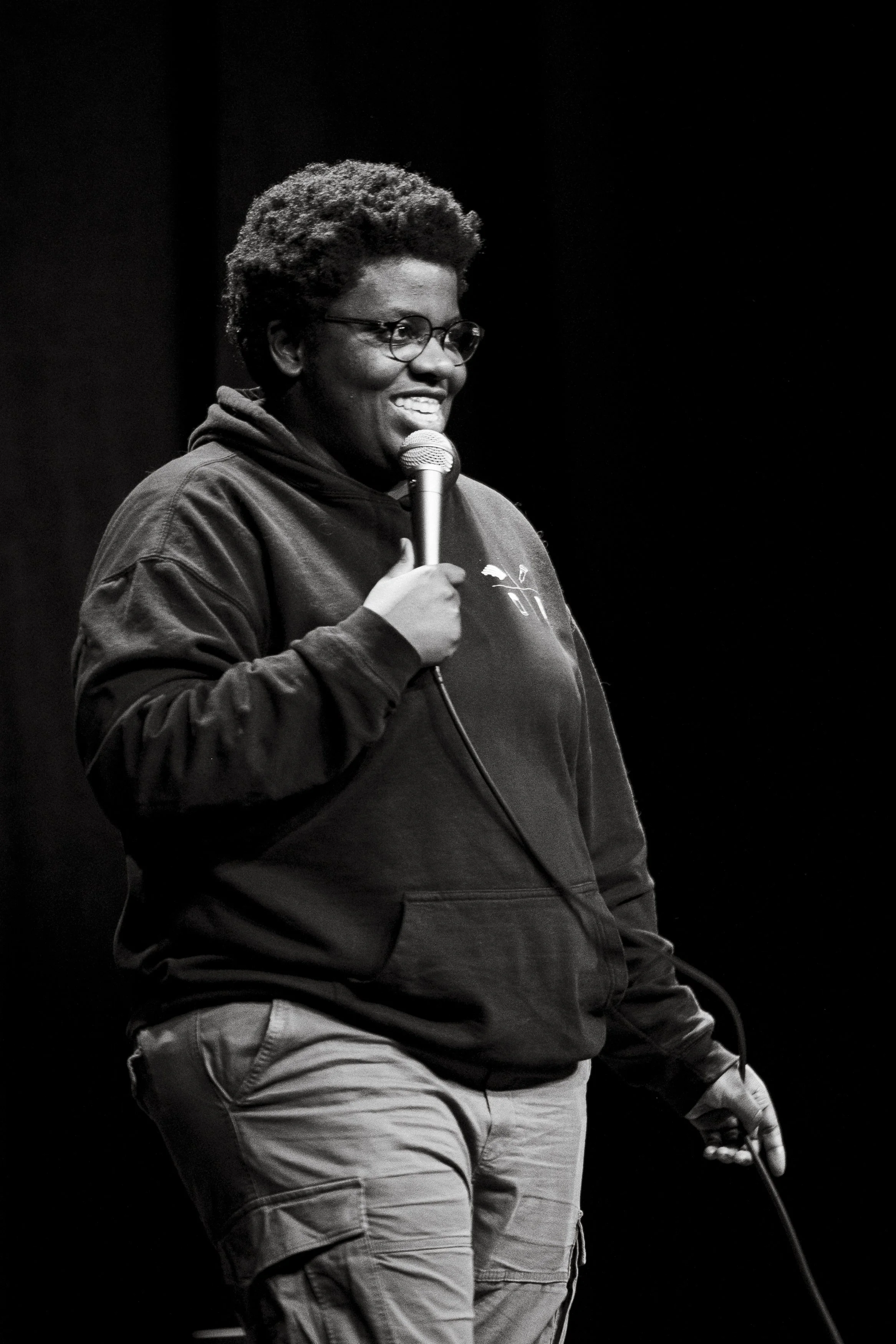 En svartvit bild av en person som talar i mikrofon, bär glasögon, hoodie och cargo-byxor, står mot en mörk bakgrund.