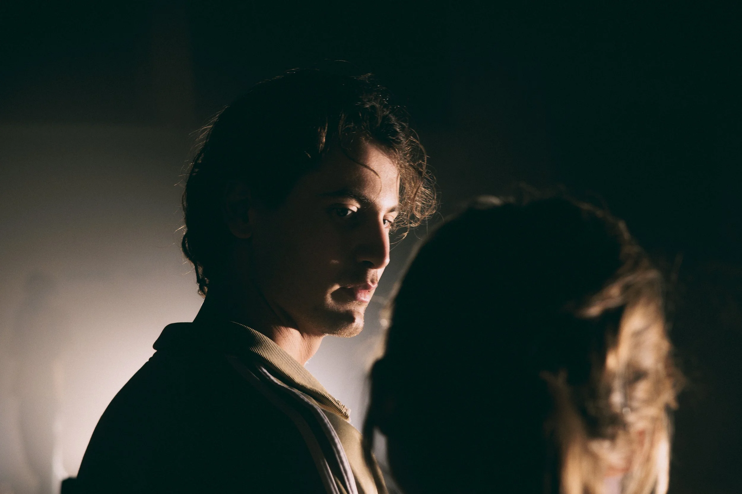 En ung man med lockigt hår tittar tillbaka mot kameran, är i skuggor med mjukt ljus som lyser upp hans ansikte.