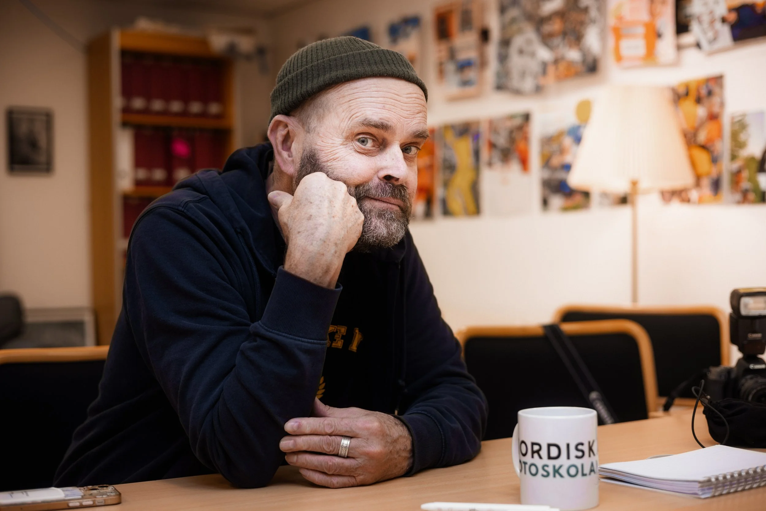 En äldre man med grått skägg och mustasch, bär en mörk hoodie och en grön mössa, sitter vid ett bord i ett rum med konst på väggarna, en skrivbordslampa och ett vitt kaffekrus med text.