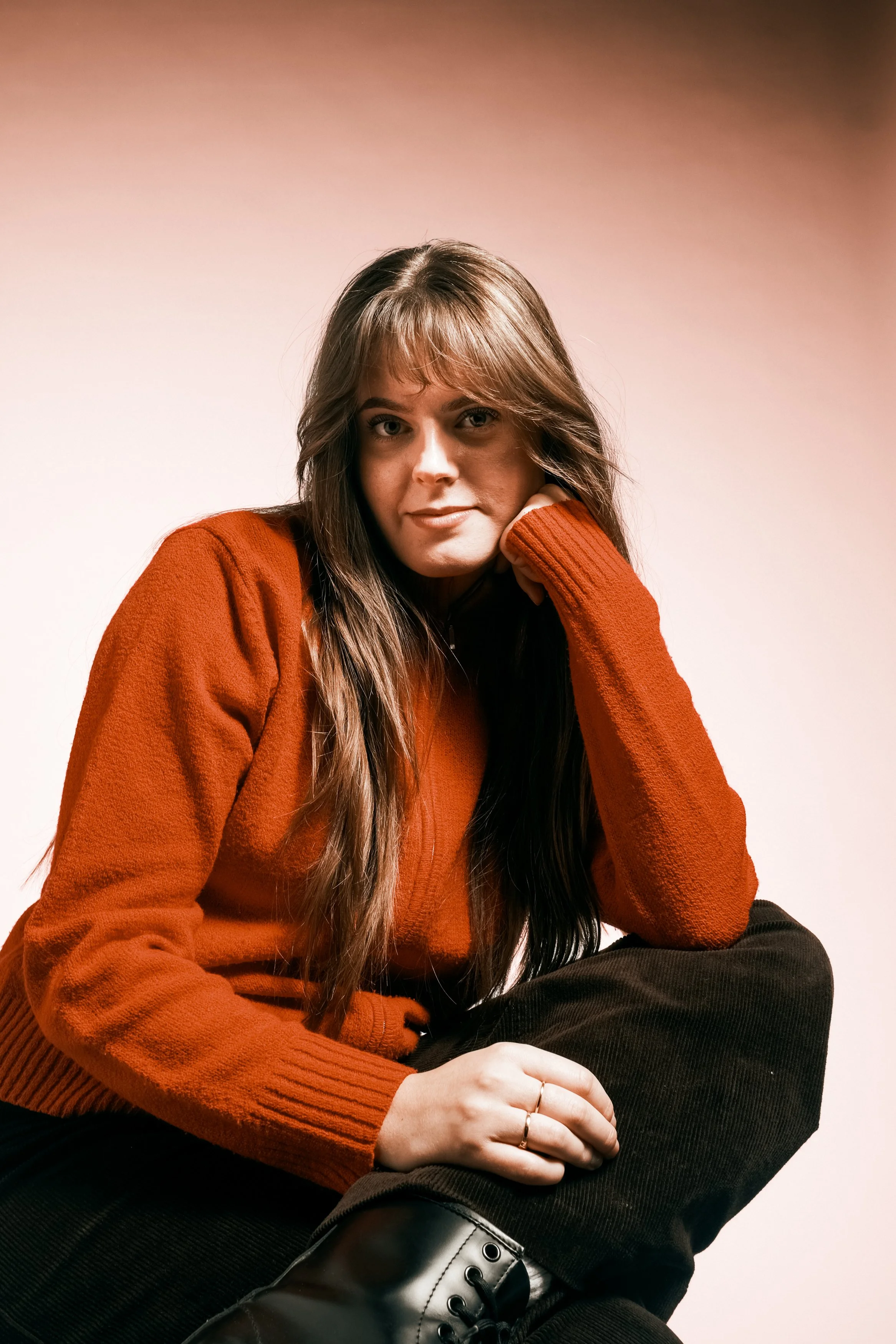 En ung kvinna med långt brunt hår, bär en orange tröja och svart byxor, sittande och vilar sitt huvud på sin hand.