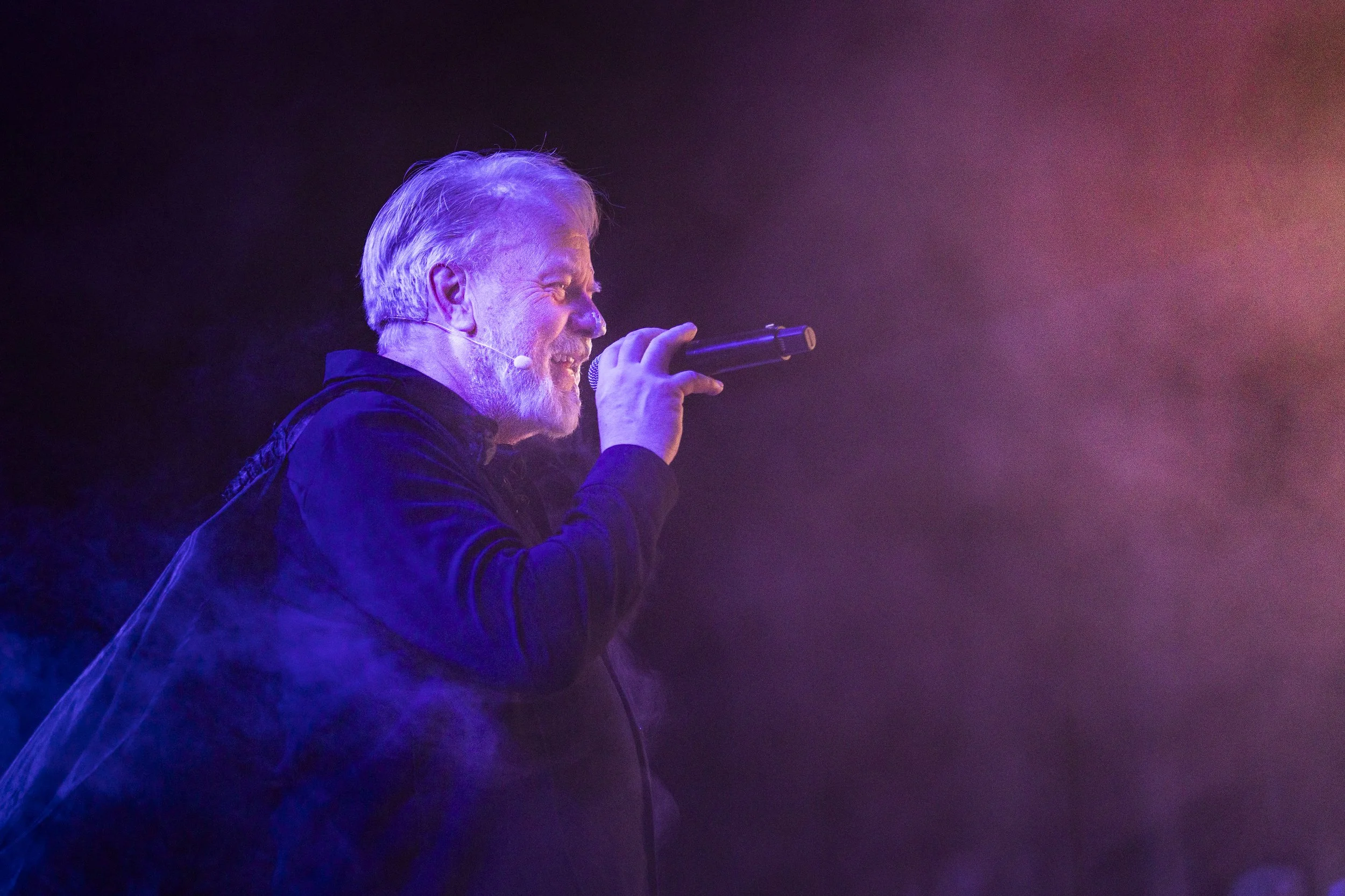En man med vit hår och skägg som sjunger i en mikrofon på scen, omgiven av färgad scenbelysning och rök.