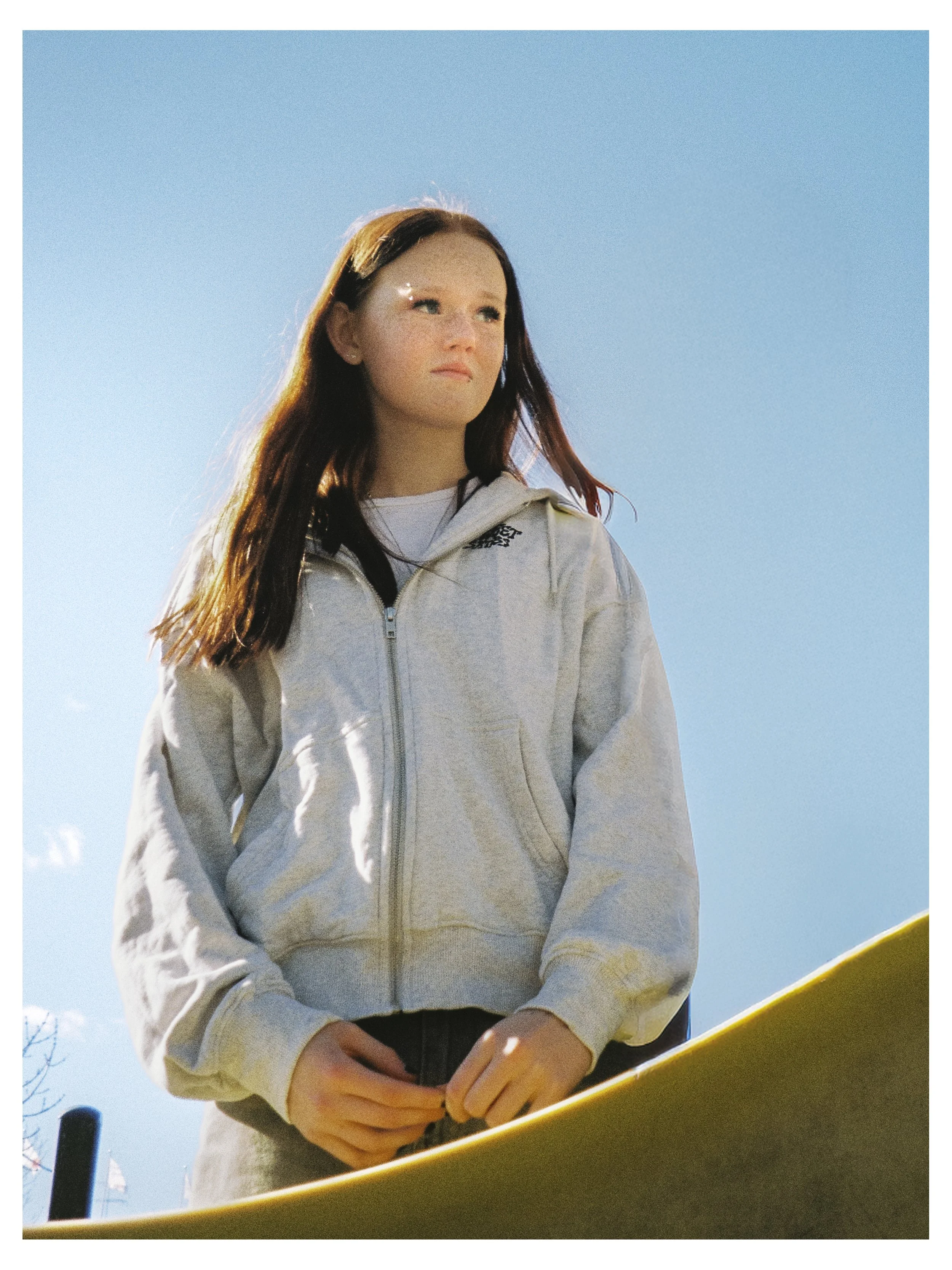 En ung kvinna står utomhus med klarblå himmel i bakgrunden, hon bär en ljus hoodie och ser tänkfull ut.