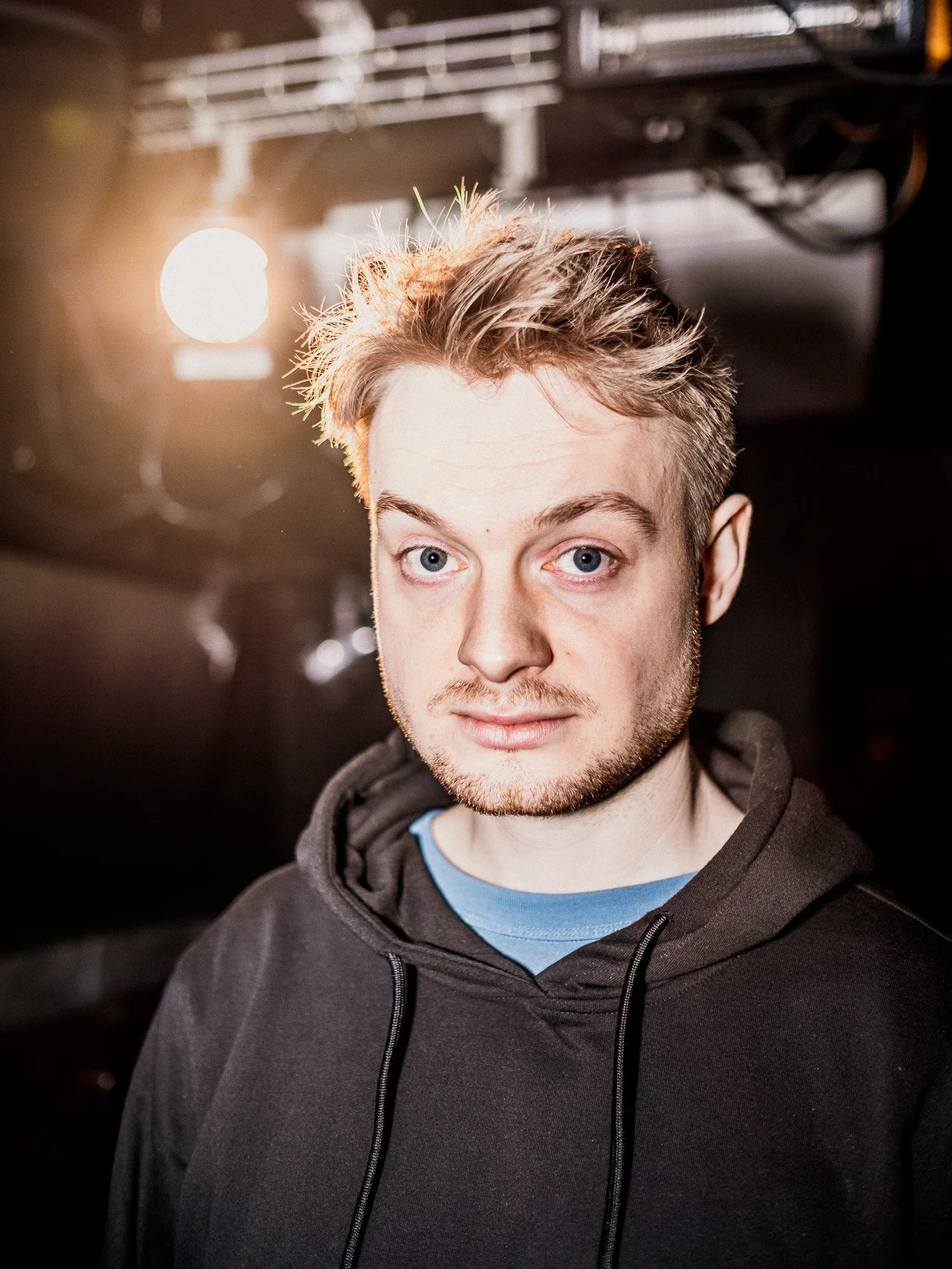 En ung man med blont, lite rufsig hår och blå ögon, bär en svart hoodie och en blå T-shirt, står i ett mörkt rum med en stark ljuskälla bakom honom.