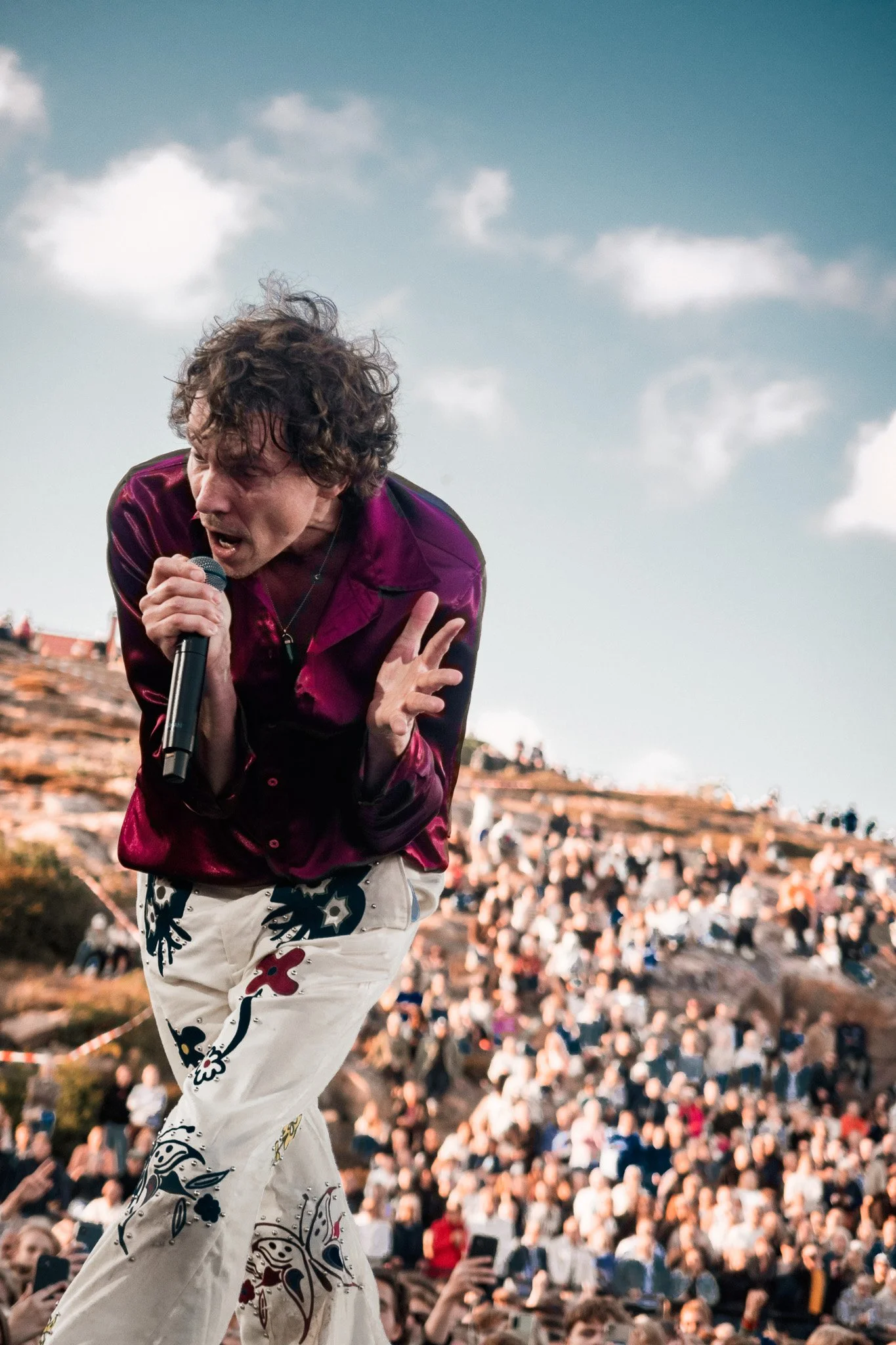 En man med mörkt, lockigt hår som sjunger eller talar in i en mikrofon på scen, med ett stort publik i bakgrunden.