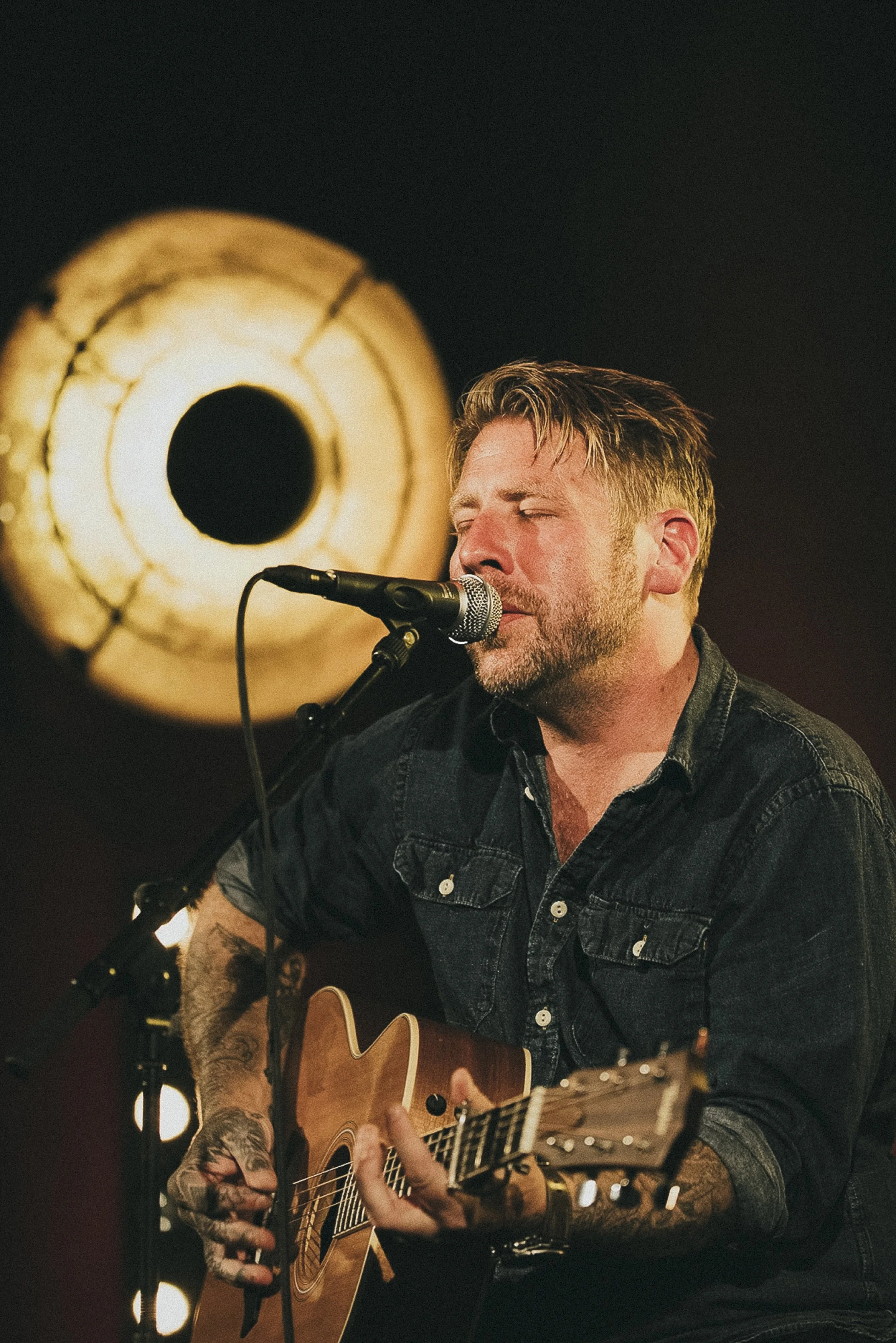Man som spelar gitarr och sjunger i mikrofon i ett mörkt rum med ett stort, varmt ljus i bakgrunden.