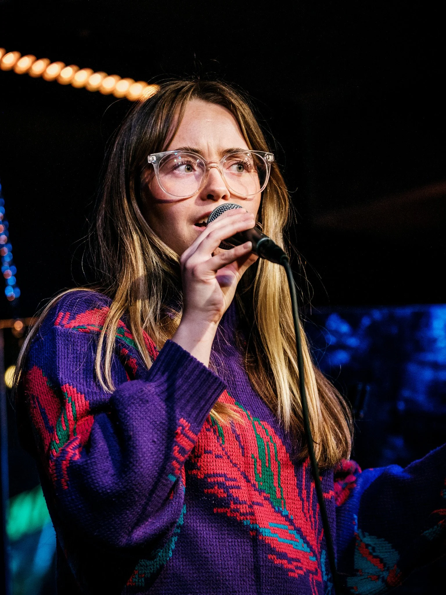 Kvinna med glasögon som sjunger eller talar i en mikrofon, iklädd en färgglad, mönstrad jacka, i en mörk miljö med några ljusa belysningar.