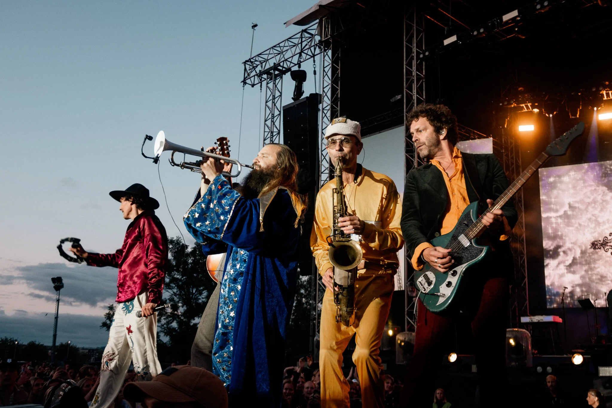Fyraklädda musiker spelar på scen på en konsert, med ett stort utomhusark och publik i bakgrunden.