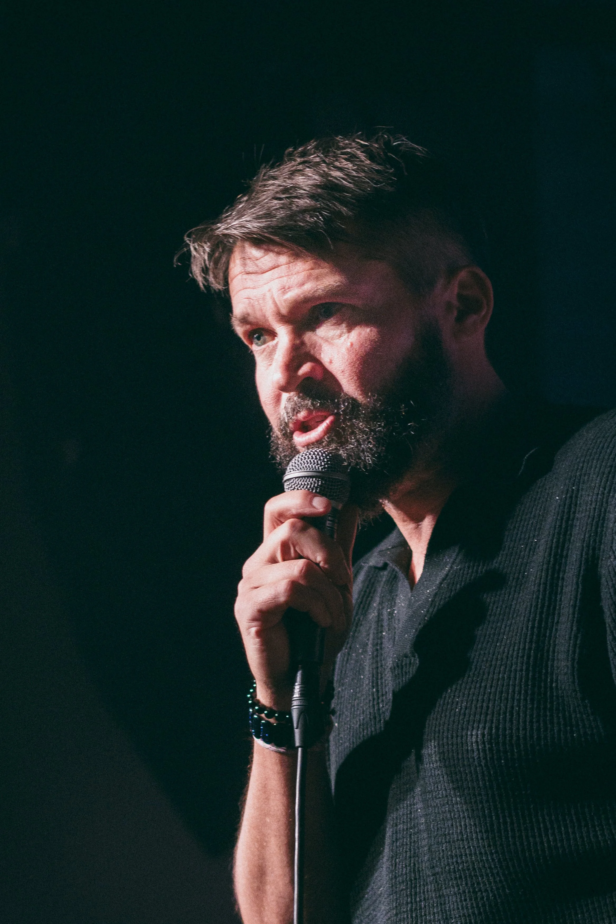 En man med mörkt hår och skägg håller en mikrofon och talar på scenen, bakgrund är mörk.