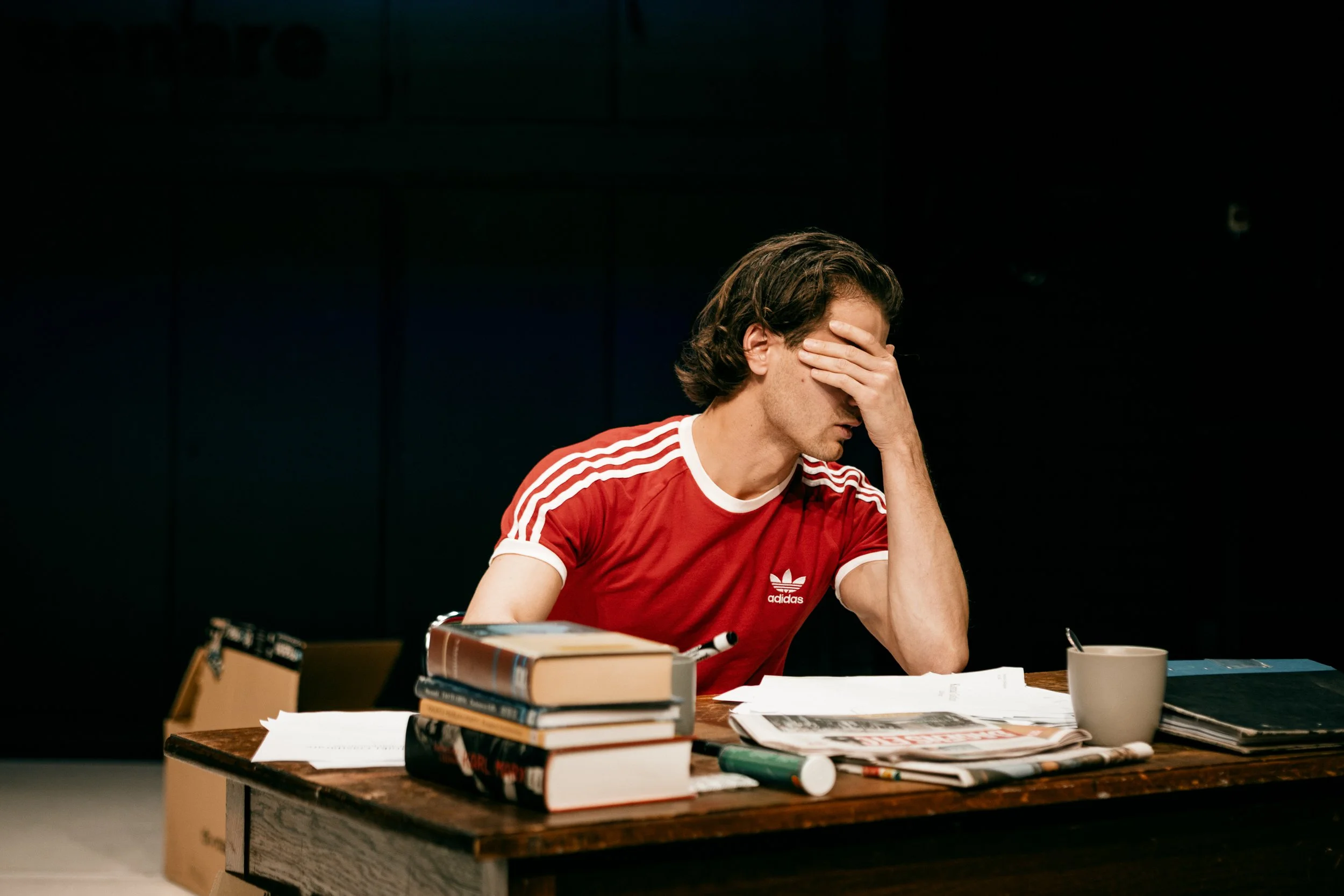 En man i en röd Adidas-tröja sitter vid ett skrivbord fullt med böcker, papper och en mugg, och ser bekymrad ut med handen mot ansiktet.