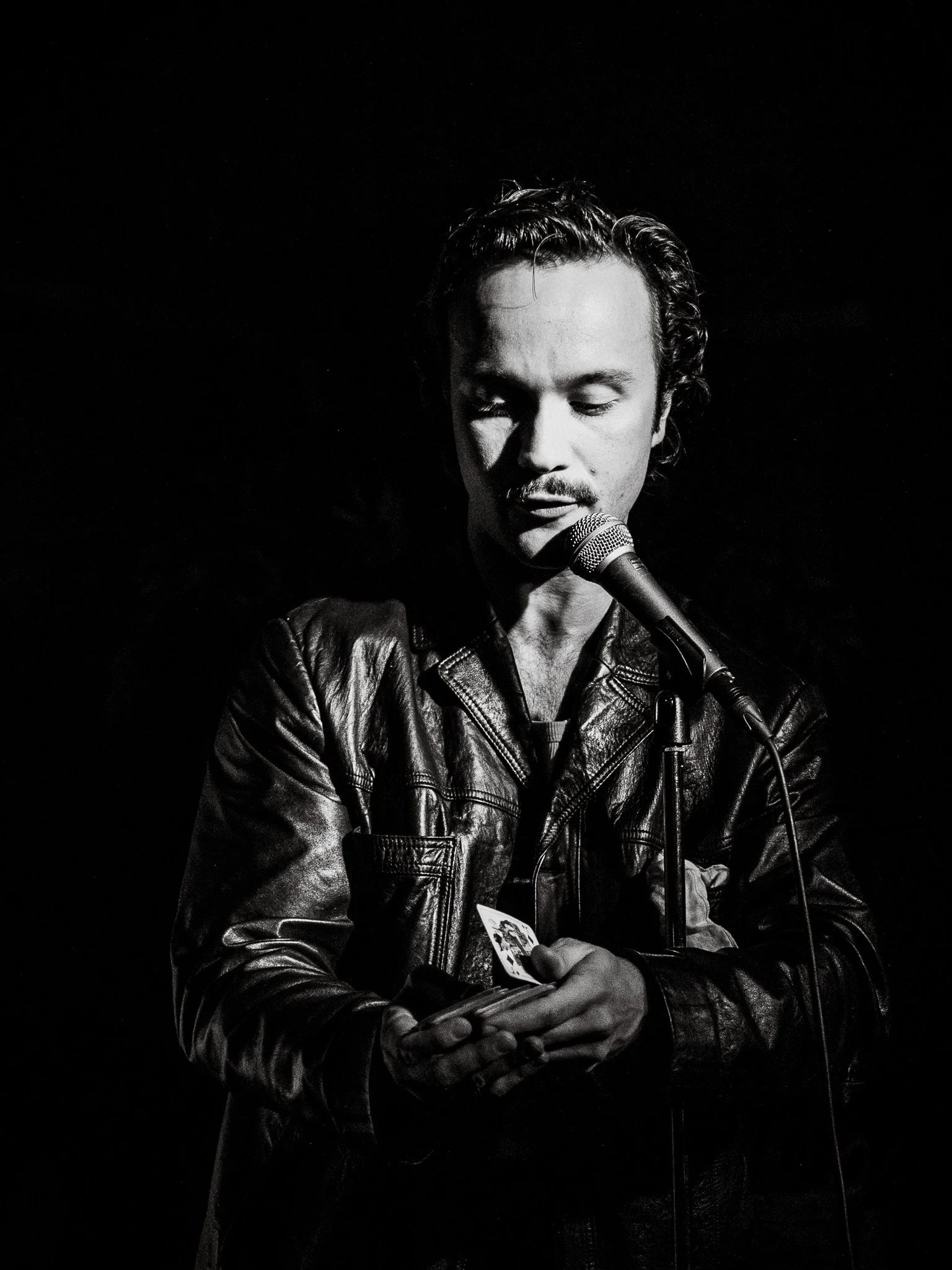 En man i läderjacka som står framför en mikrofon och håller ett kort i handen, med ett set som är mörkt och en svart bakgrund.