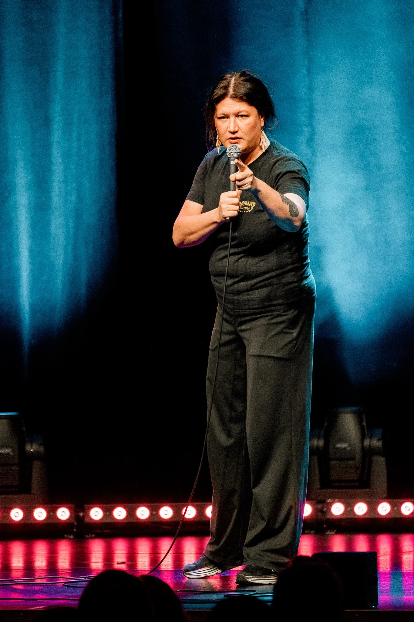 Kvinna som står på scen med mikrofon, riktar sig mot publiken, bakgrund med blåa och svarta kulisser.