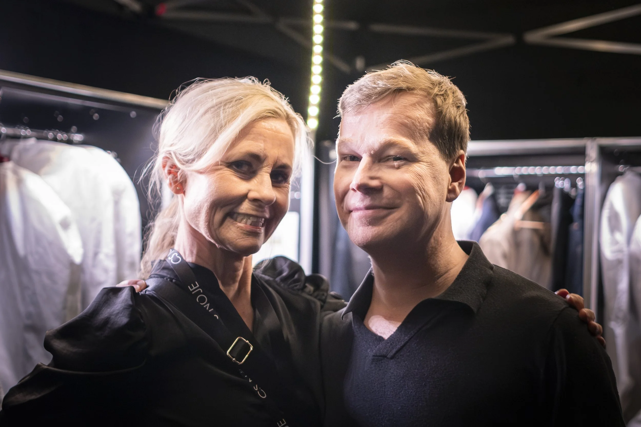 En kvinna och en man som ler och står nära varandra i en klädbutik eller backstage, med kläder på galgar i bakgrunden.