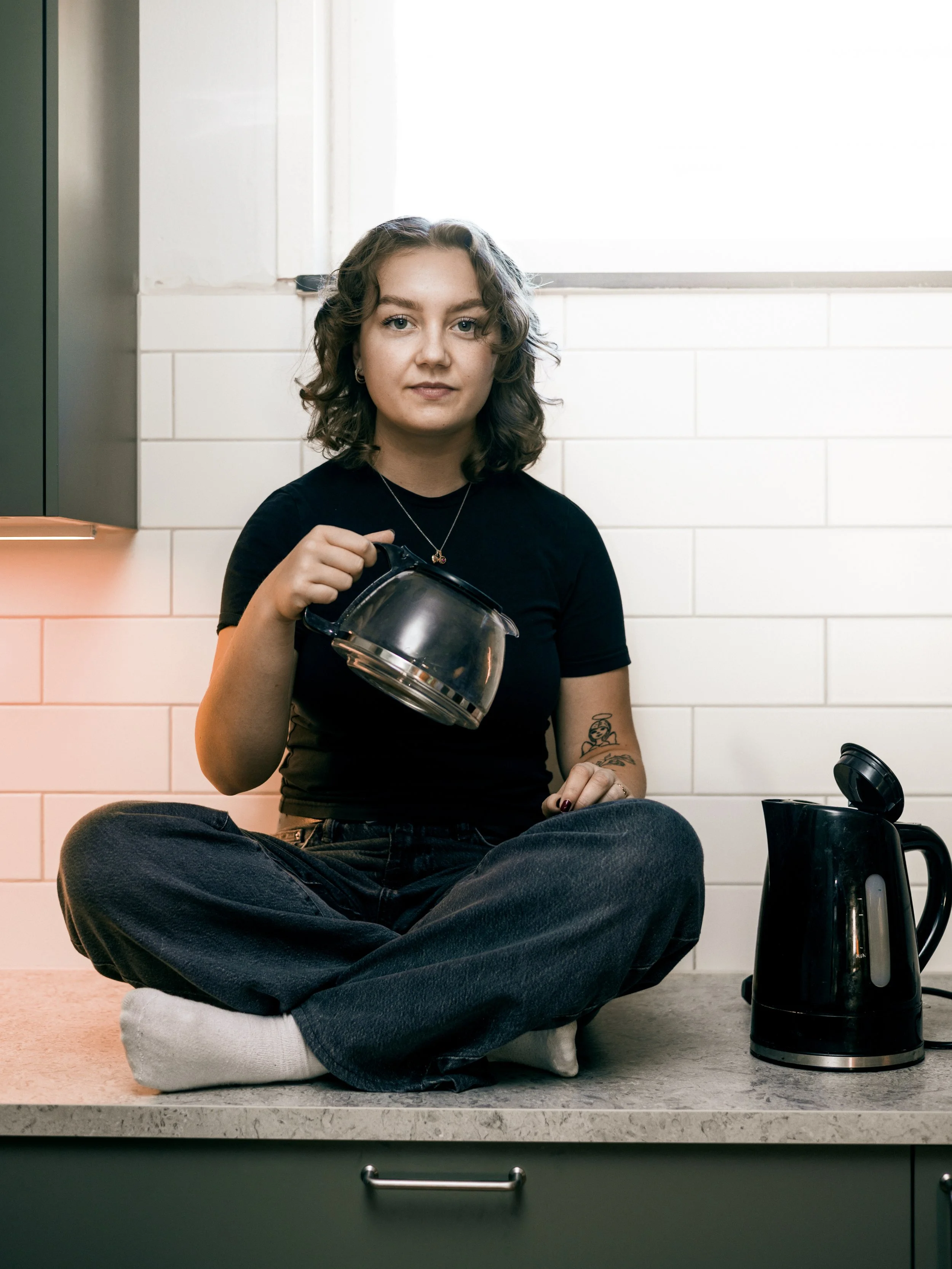 Ung kvinna med lockigt hår, klädd i svart, sitter på köksbänken med korslagda ben och rör en glaskanna, med el-kokare på köksbänken.