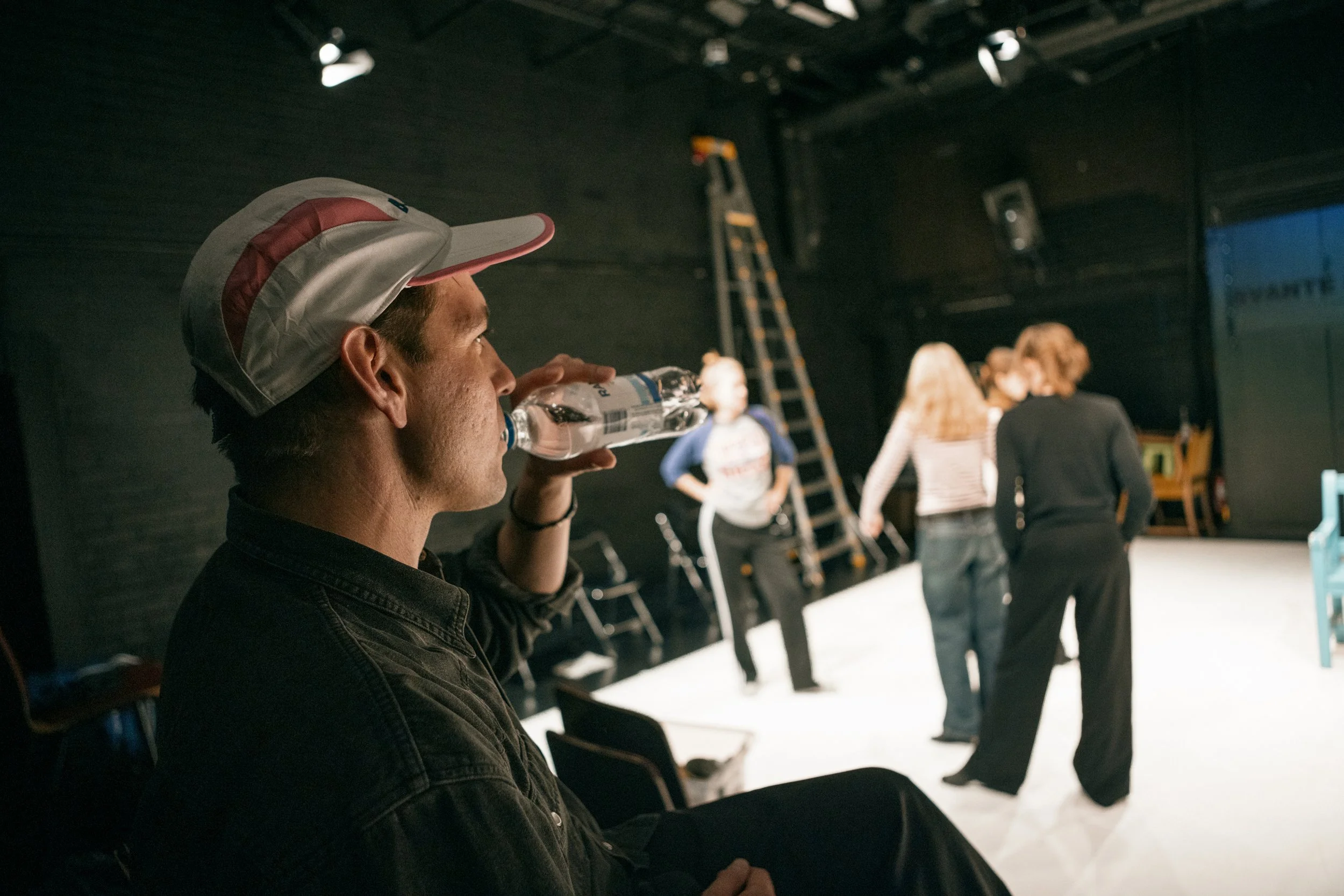 En man i profil som dricker vatten medan han sitter på en svart stol, med en grupp människor som pratar och står i en cirkel i bakgrunden i ett mörkt rum.